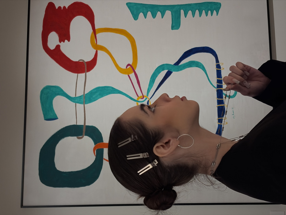 The image displays a close-up profile shot of a young person positioned in front of a vibrant, abstract painting. The subject, likely female, is looking slightly upwards and to the left, with her head tilted back. Her dark hair is neatly pulled back into a low bun, and several silver duckbill hair clips are visible on the side of her head. She wears a large, simple silver hoop earring in her left ear. Around her neck are multiple necklaces; one, a delicate chain with small dark beads, is held lightly between the fingers of her right hand, which extends into the frame from the right side. Her left shoulder and arm, covered by a black garment, are also visible.
The background is a large, predominantly white abstract painting. It features an array of colorful, free-form shapes and wavy lines that appear to emanate from and connect to each other. Key elements in the painting include:
* A large, irregular red shape with an internal cut-out on the upper left.
* A thick yellow wavy line that curves upwards and then forms a loop, connecting to other elements.
* Multiple teal (light blue-green) wavy lines, some forming loose loops, one of which has a thinner brown loop hanging from it.
* A prominent dark blue wavy line, which also extends towards the right.
* A thin magenta line that seems to connect the yellow line to the area around the subject's eye.
* A comb-like or jagged teal shape at the top center.
* A large, thick dark green oval-like shape on the lower left.
* An orange C-shaped element on the lower left, near the dark green shape.
The composition suggests an artistic interpretation of thoughts or energy flowing from the person's mind, as several lines and shapes in the painting appear to originate from or converge towards her eye level. The lighting is soft and even, highlighting the subject's features and the colors of the painting. Faint, illegible text or a signature, possibly "Emy" and a number like "99", is barely visible on the painting towards the right side. The overall mood is contemplative and artistic.