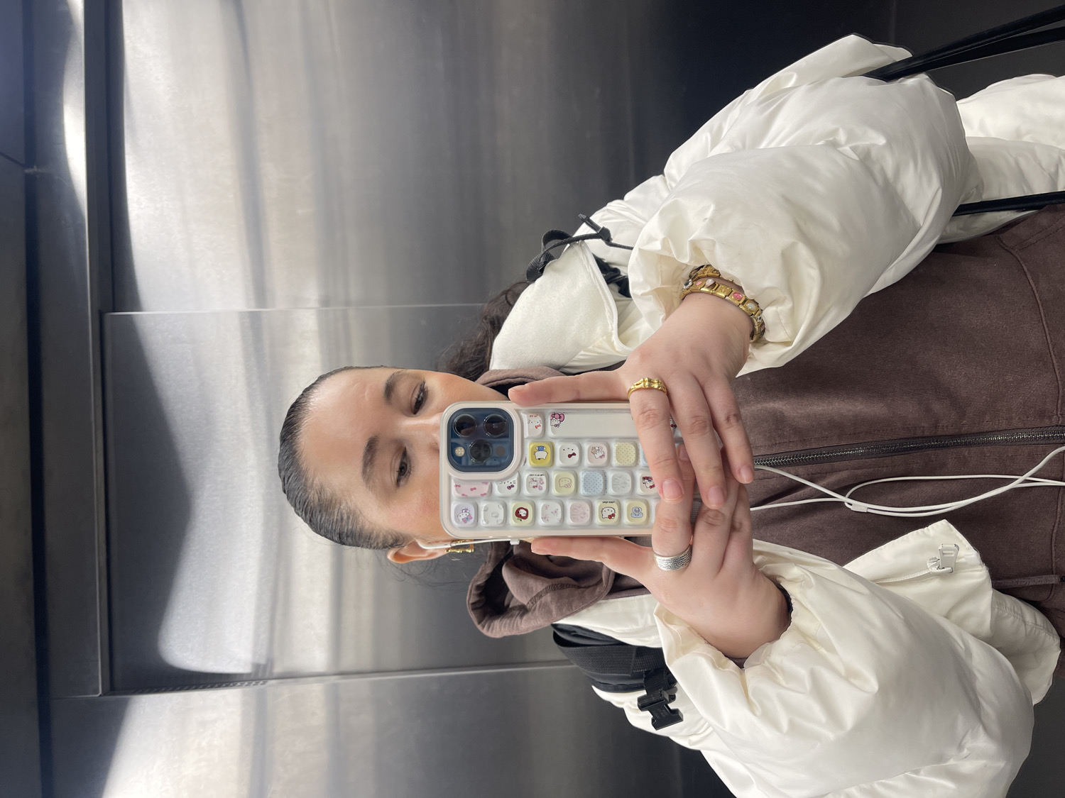 An upward-oriented mirror selfie captures a young adult woman inside what appears to be an elevator in Moscow, Russia. She is shown from the chest up, with her dark hair neatly pulled back. Her attire consists of a white, voluminous puffy jacket worn over a brown hooded jacket or sweatshirt, which features a zippered front and a soft texture, possibly velour.
The woman is holding a white iPhone with a distinctive light-colored case adorned with multiple small, colorful Sanrio characters, including Hello Kitty. Her attention is focused on the phone screen as she takes the picture. She accessorizes with several pieces of jewelry: a gold-toned, intricately designed ring on her right hand, and multiple bracelets on her right wrist, one of which has gold links and pinkish accents. On her left hand, she wears a silver-toned ring with a textured, possibly ribbed pattern. Small gold hoop earrings are also visible.
The background is composed of reflective, brushed metallic panels, typical of an elevator interior, featuring horizontal seams. The indoor setting implies artificial lighting. No discernible text is present on her clothing or the background, although the phone case prominently features character designs.
