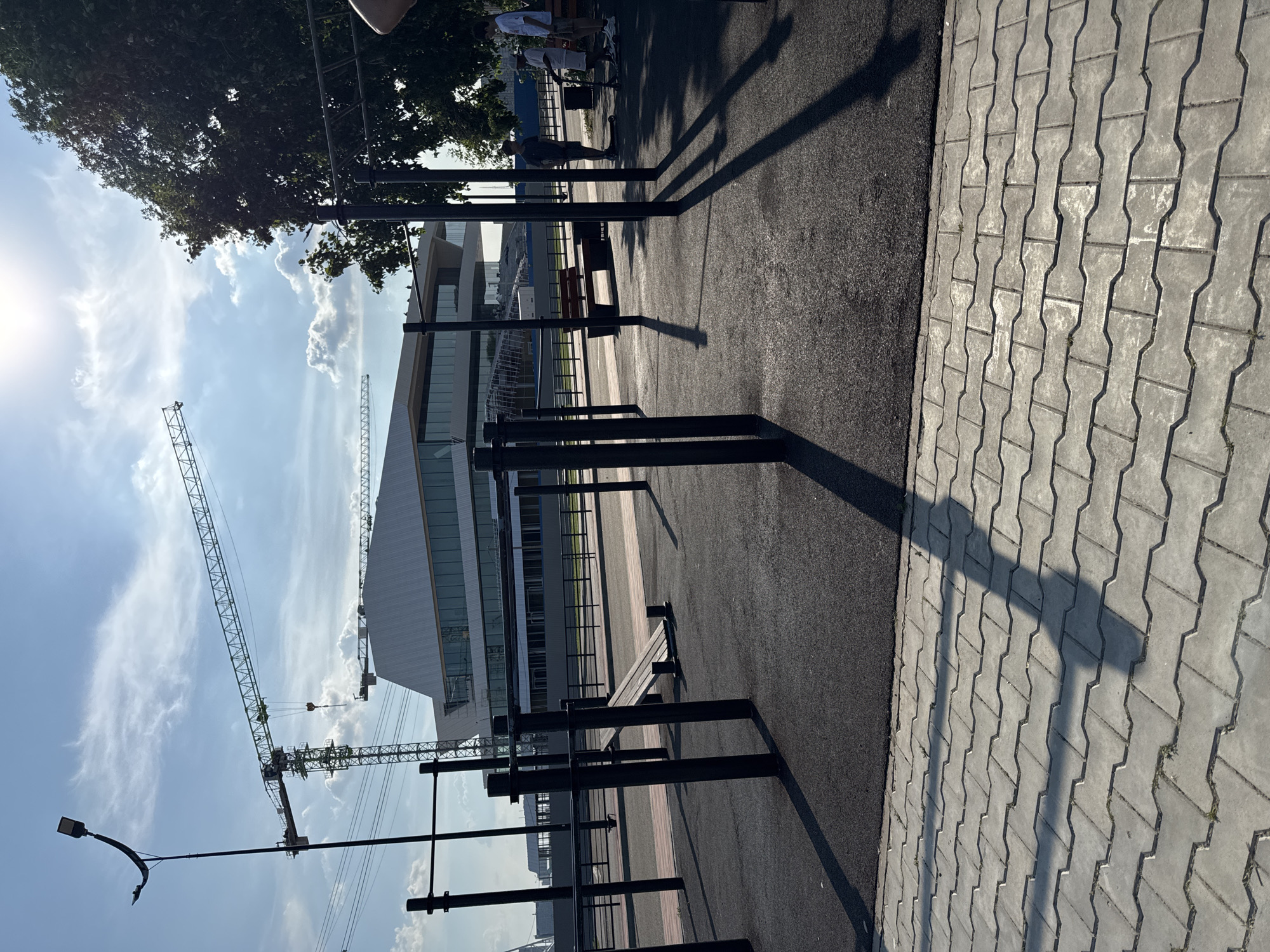 The image depicts an outdoor exercise park in Blagoveshchensk, Russia. In the foreground, there are black metal bars for calisthenics, set on a dark rubberized surface. Beyond the park, a paved walkway made of grey interlocking bricks is visible, with long shadows cast by the exercise equipment. In the middle ground, a bench sits near the exercise area. Further back, a modern, multi-story building with a distinctive white, angular roof and large glass windows is under construction. Two construction cranes are positioned near the building, one large tower crane and another smaller one. A street light with a curved arm is also visible in the background. The sky is blue with scattered white clouds, and the sun is shining brightly. Three young males are present in the image. One is standing on a scooter, another is standing next to him, and a third is standing slightly further away. They are all dressed casually. The overall scene suggests an urban outdoor recreational space undergoing development.