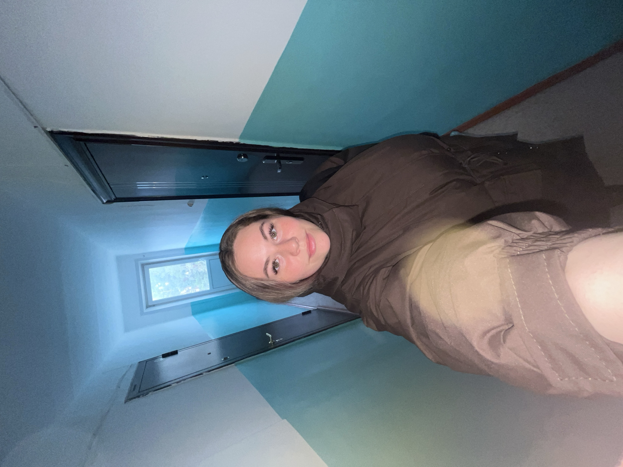 The image, taken in Chaikovsky, Russia, shows a young woman capturing a selfie in what appears to be a narrow hallway or stairwell. The woman, with light brown hair and green eyes, looks directly at the camera with a neutral to slightly pleasant expression. She is wearing a dark brown jacket or hoodie and light-colored pants.
The setting is characterized by walls painted in two distinct horizontal sections: the lower portion is white, and the upper portion is a light blue or turquoise. Dark-colored, possibly metal, doors are recessed into the walls on both sides of the hallway, likely serving as entrances to apartments or utility access points. At the far end of the hallway, a small rectangular window lets in natural light, through which green foliage is visible, suggesting an outdoor view. The ceiling is white.
The soft, natural lighting from the window indicates that the picture was taken during the daytime. No text is visible in the image.