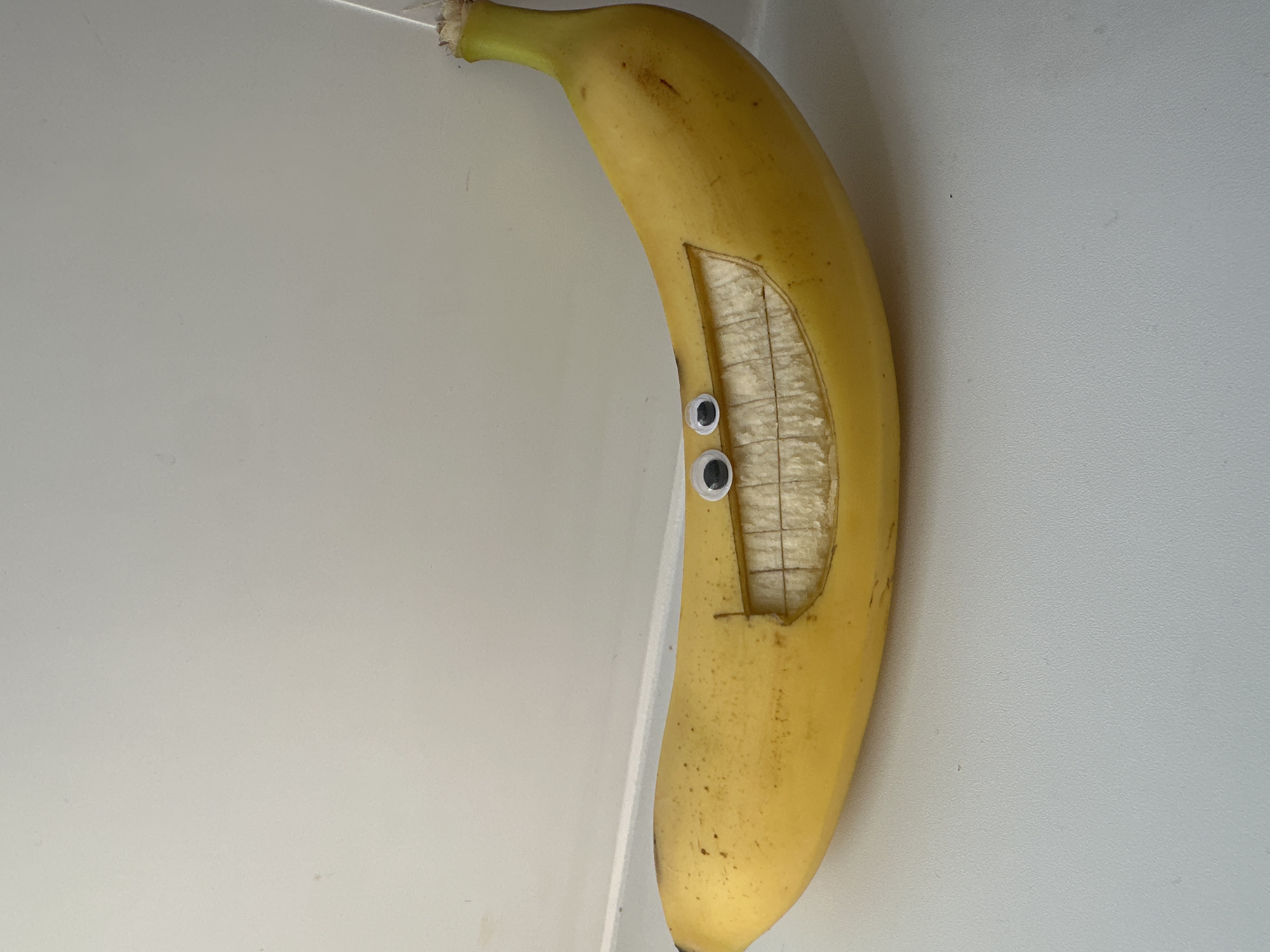 A ripe, yellow banana is prominently displayed against a plain, light gray or off-white background, possibly a wall or table surface. The banana has been creatively transformed into a character, featuring two googly eyes adhered to its skin. Below the googly eyes, a rectangular section of the banana peel has been cut away, revealing the pale inner fruit. This exposed area functions as a "mouth," and faint, shallow horizontal and vertical etchings on the banana's flesh within this opening give the impression of teeth or a textured interior. The banana's stem is visible at the top left of the image. The googly eyes and carved mouth give the banana a cheerful, surprised, or comical expression. The lighting is even, suggesting an indoor setting. No specific time of day or weather conditions are discernible, and there is no visible text in the image. This media file originates from Votkinsk, Russia.