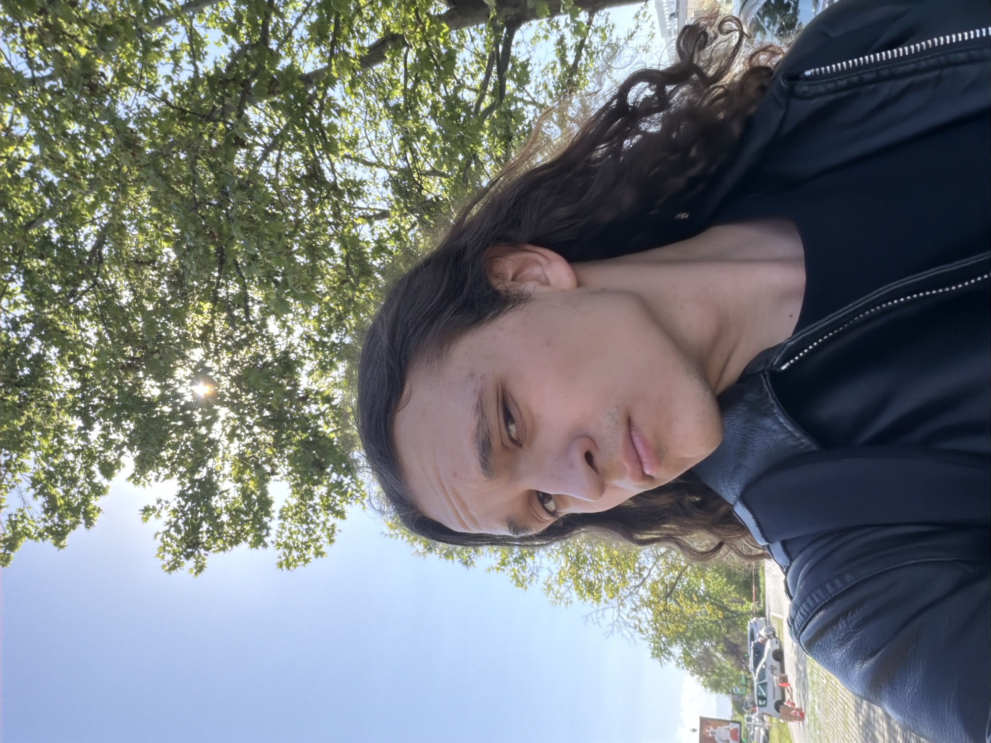 The image is a tilted, close-up portrait of a person with long, dark, wavy hair and fair skin. They are seen from a low angle, wearing a black jacket, possibly leather, over a black top. The person has a neutral to slightly contemplative expression, looking towards the camera. Notable details include visible wrinkles on the forehead and minor skin imperfections.
The background depicts an outdoor scene on a bright, sunny day. The clear blue sky occupies the lower portion of the frame, with the sun brightly shining and peeking through the abundant green leaves of trees overhead. In the distance, a paved area, which could be a street or parking lot, is visible, along with several parked vehicles, including a white van or truck. A blurry white sign featuring what appears to be a white cat and some indistinct text, along with a smaller red sign, are visible in the lower right background. The individual appears to be taking a selfie in this outdoor setting, likely in Plovdiv, Bulgaria.