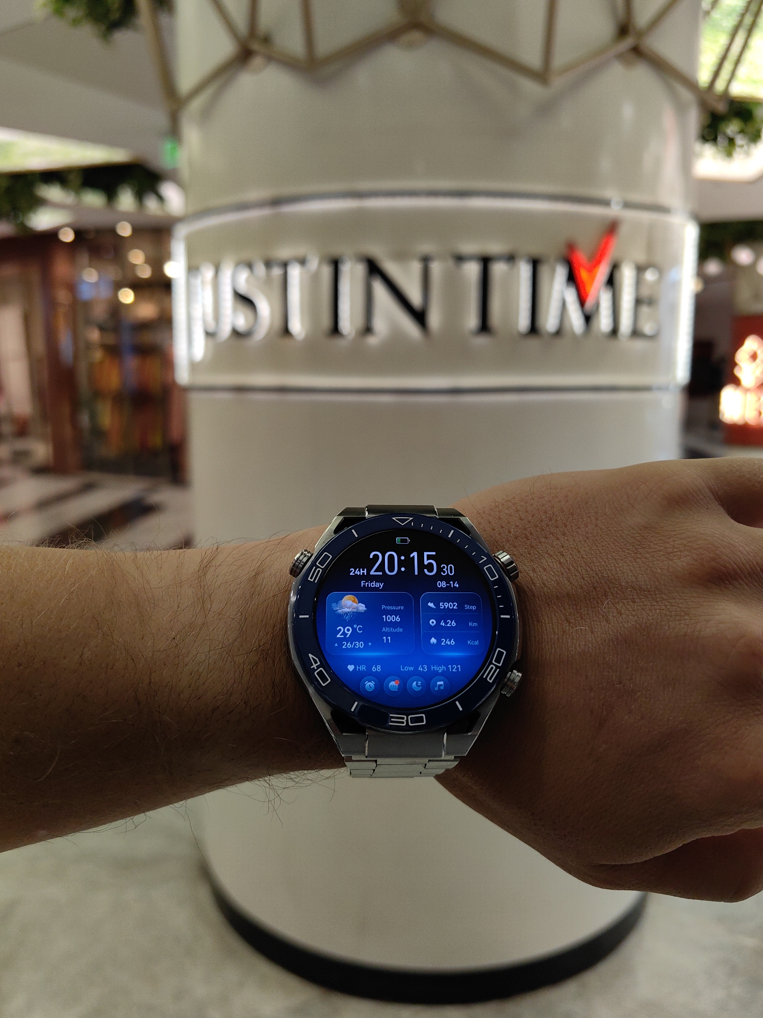 A close-up shot shows a person's wrist wearing a smartwatch with a metallic band. The smartwatch displays the time as 20:15:30, the date as 08-14, and various health and weather metrics, including a temperature of 29°C, 5902 steps, and a heart rate of 68. In the blurred background, a large, white cylindrical structure is visible, adorned with illuminated text that reads "JUSTIN TIME." The environment appears to be an indoor public space, possibly a mall or a commercial establishment, with a modern architectural style. The lighting suggests an artificial interior setting. No specific location in Mumbai, India, can be identified from the visual information provided.