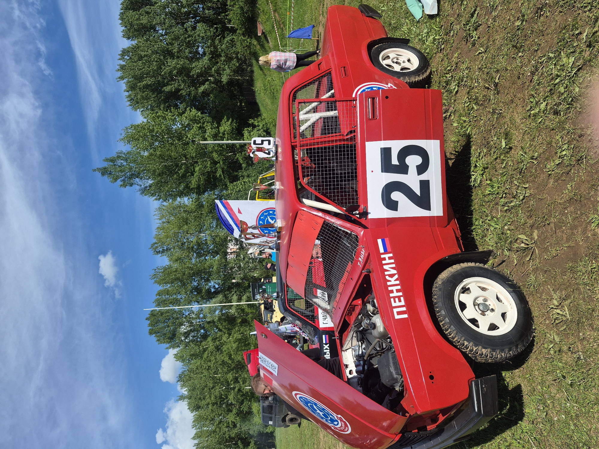 A bright red modified race car labeled "25" is parked on a grassy field under a partly cloudy sky in Unknown, Unknown. The hood of the car is open, revealing the engine and a person working on it. Additional people are visible in the background near the car. Trees are also in the background.