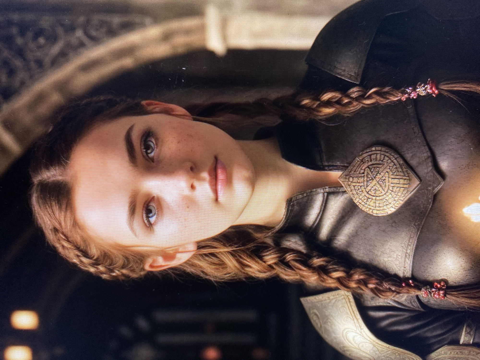 A close-up, head and shoulders portrait of a young woman with auburn hair braided into two pigtails adorned with red beads. She has fair skin with freckles, striking blue eyes, and is wearing a dark leather outfit with ornate gold pauldrons and a large, circular, gold pendant with intricate carvings. The background is dark and appears to be an interior with architectural elements and soft, out-of-focus lighting. The scene is set in the city of Unknown, Unknown.