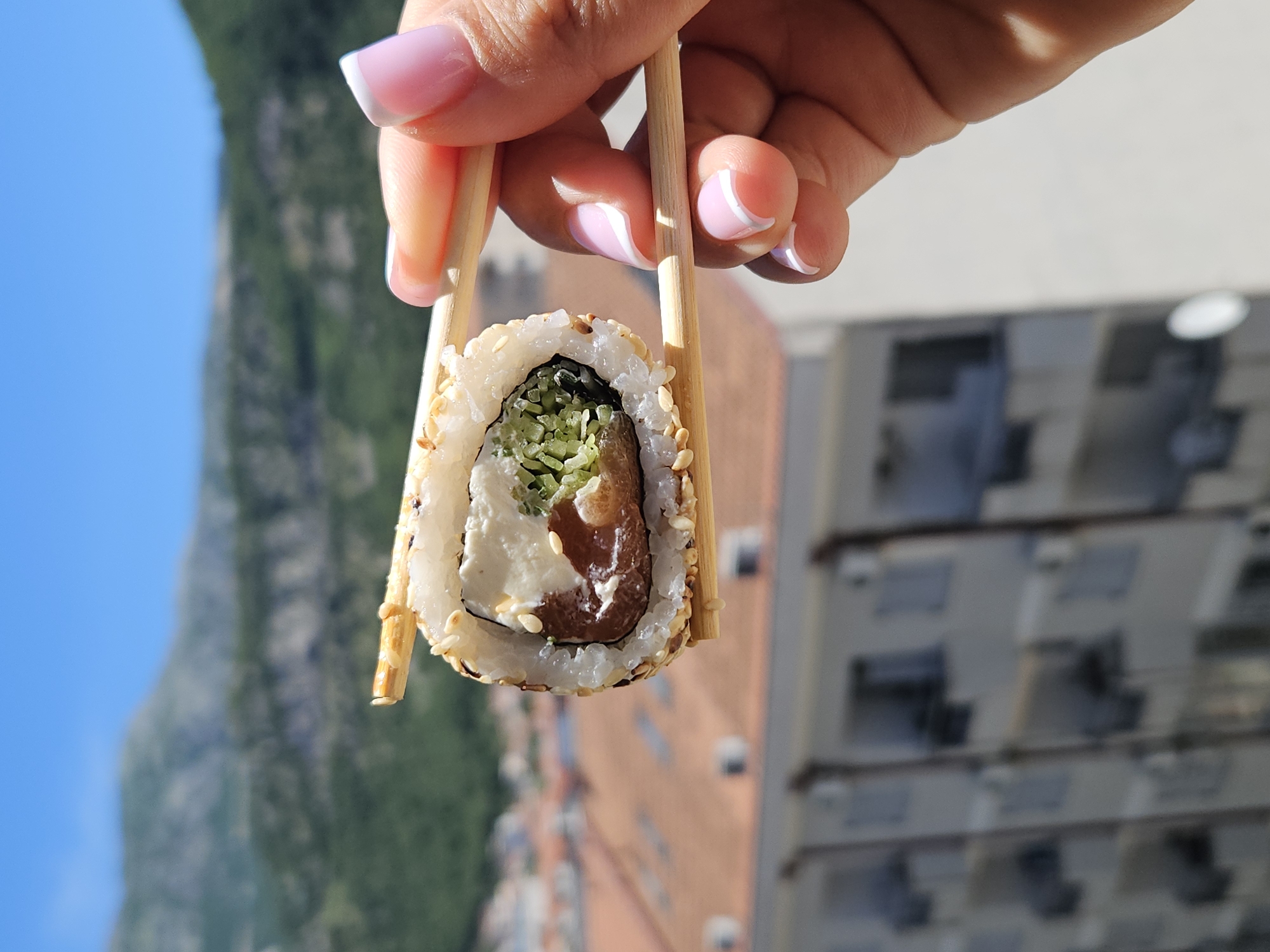 A hand with pink and white manicured nails holds a piece of sushi with chopsticks. The sushi is covered in sesame seeds and contains what appears to be rice, a white creamy filling, a dark-colored ingredient, and chopped greens. The background shows a building with windows and a mountain range against a blue sky. The city is unknown.