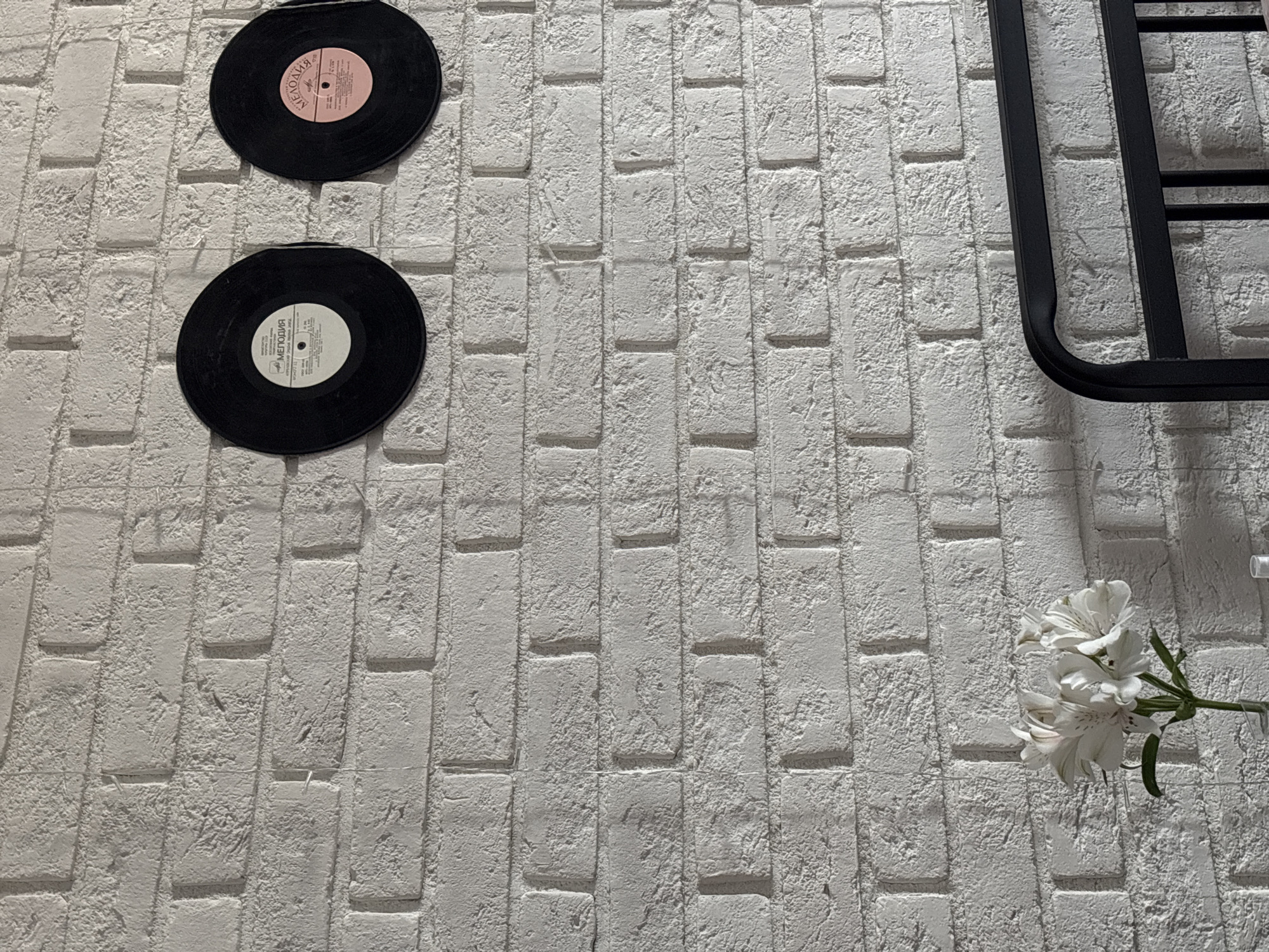 A high-angle shot depicts a white brick wall adorned with two vinyl records on the left side. One record features a pink label, while the other has a white label. To the right, a black metal frame is partially visible, and a bouquet of white alstroemeria flowers is placed near the lower right corner of the frame. The scene is set against a white brick wall in what appears to be Perm, Russia.