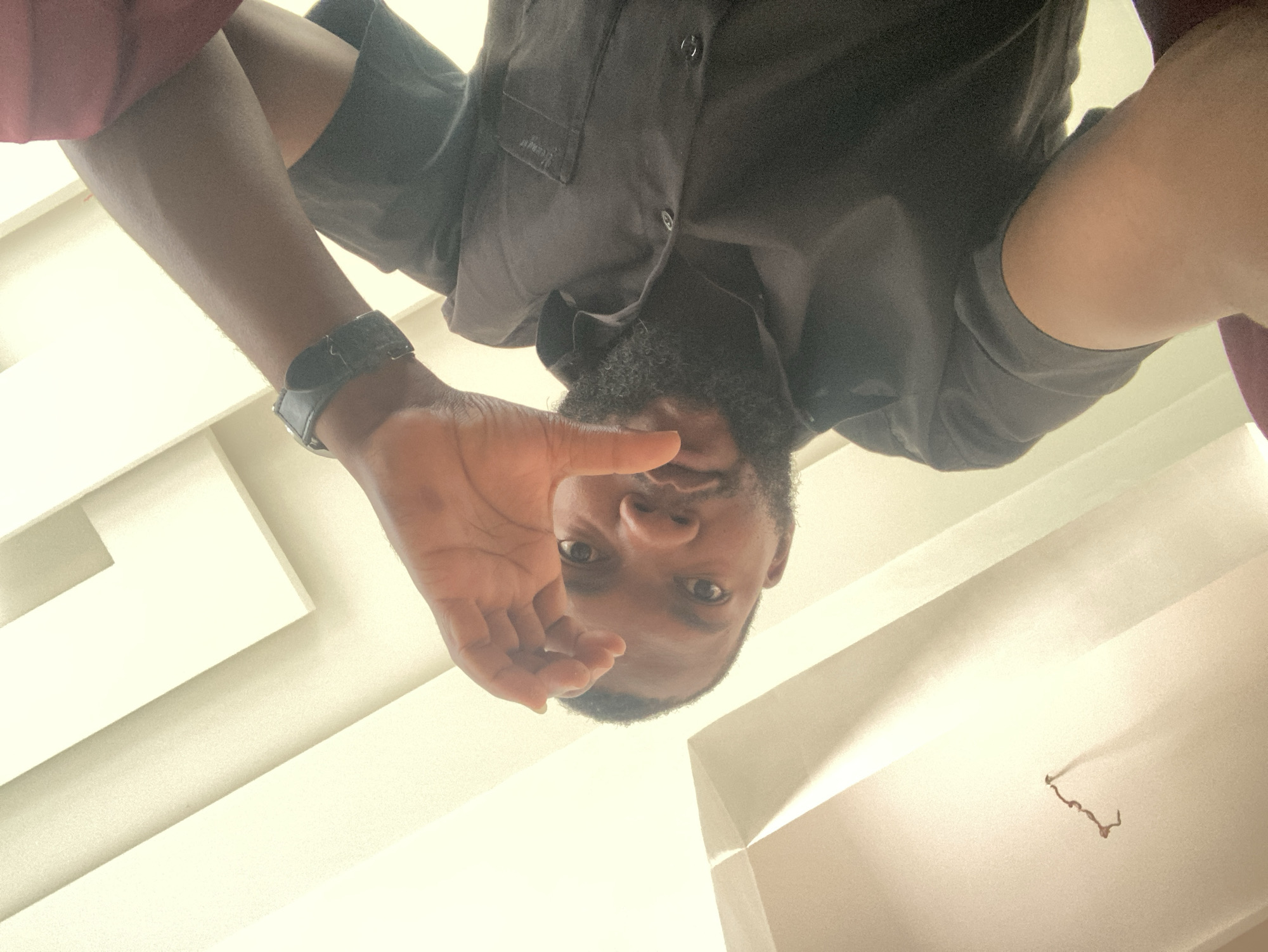 This media file captures an adult male in an indoor setting in Jos, Nigeria, from a low-angle perspective, looking directly upwards at him. The man, who has dark skin, short curly hair, and a dark beard, is positioned upside down in the frame, suggesting he is leaning over the camera. He is wearing a dark, possibly black or very dark grey, short-sleeved button-up shirt with a pocket on the left chest, where faint, illegible white text or a logo is visible. A dark watch is worn on his left wrist. His left hand is raised, with the palm slightly open, partially obscuring the lower part of his face.
The background is a brightly lit, off-white or cream-colored ceiling or wall featuring clean, geometric recessed panels or beams. In the lower right portion of the image, a dark, squiggly wire or string is seen hanging from the ceiling. The lighting is bright and diffuse, casting minimal shadows and suggesting either strong artificial light or indirect natural light. The man's expression is neutral as he looks into the camera. There is no discernible text other than the faint, illegible mark on his shirt.