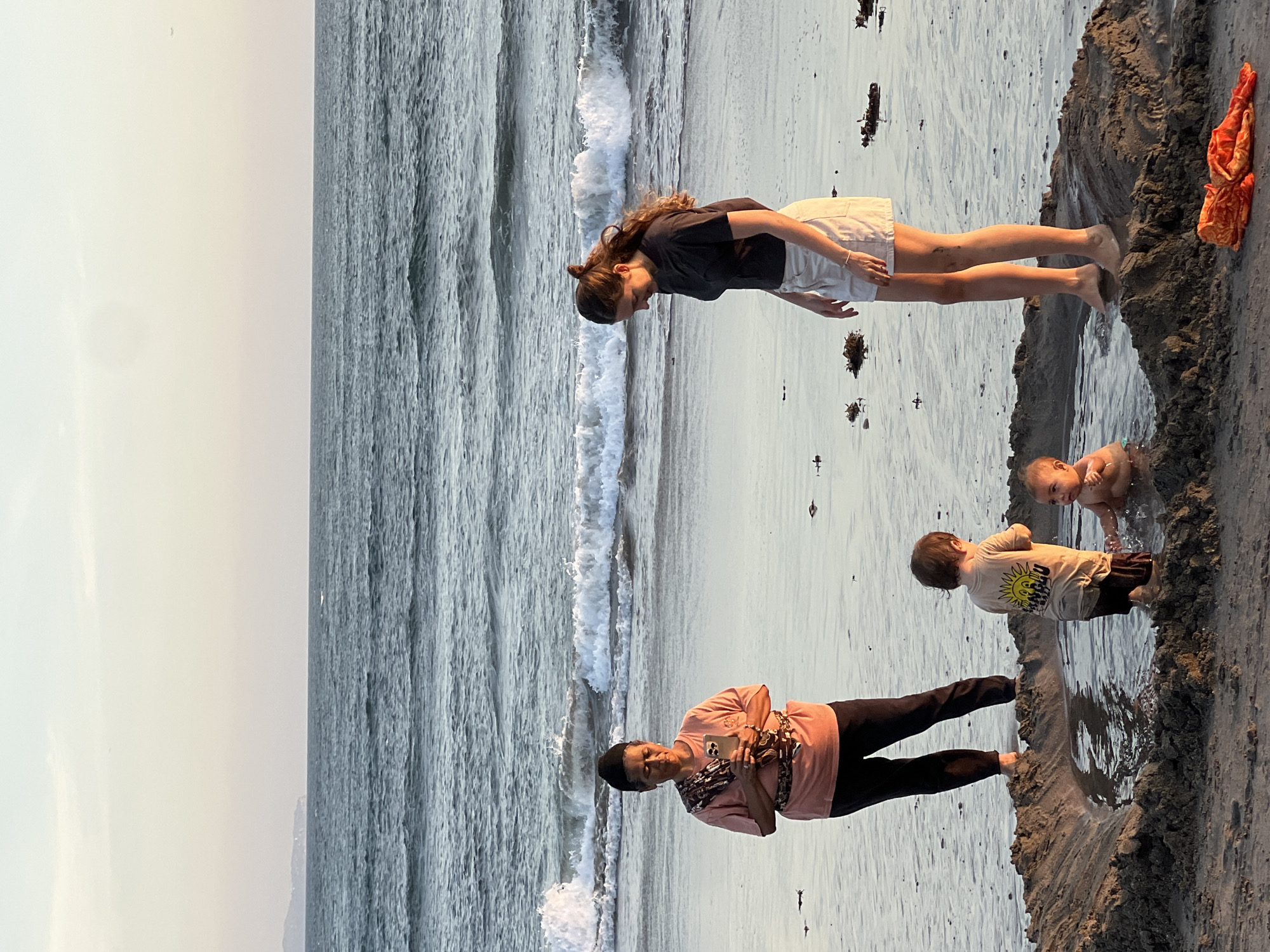 On a sandy beach in North Kuta, Indonesia, a group of four people are gathered near the shoreline. The ocean is calm with gentle waves, and the sky is a soft gradient of pale yellow and pink, suggesting either sunrise or sunset.
In the foreground, two young children are playing in a shallow, sand-lined pool. One child, wearing a light-colored t-shirt with a graphic on it, stands in the water, facing away from the camera. The other child, a baby, is seated in the water, splashing with their hands.
An adult woman, dressed in a dark t-shirt and white shorts, stands to the right of the children, looking down at them. Her feet are partially submerged in the water.
To the left, another adult, dressed in a coral-colored t-shirt and dark pants, holds a smartphone and appears to be looking at it. They are also standing near the water's edge.
The beach is wet and sandy, with scattered pieces of seaweed. A bright orange piece of fabric or clothing lies on the sand in the lower right corner of the image. In the distance, the horizon shows a faint outline of land.