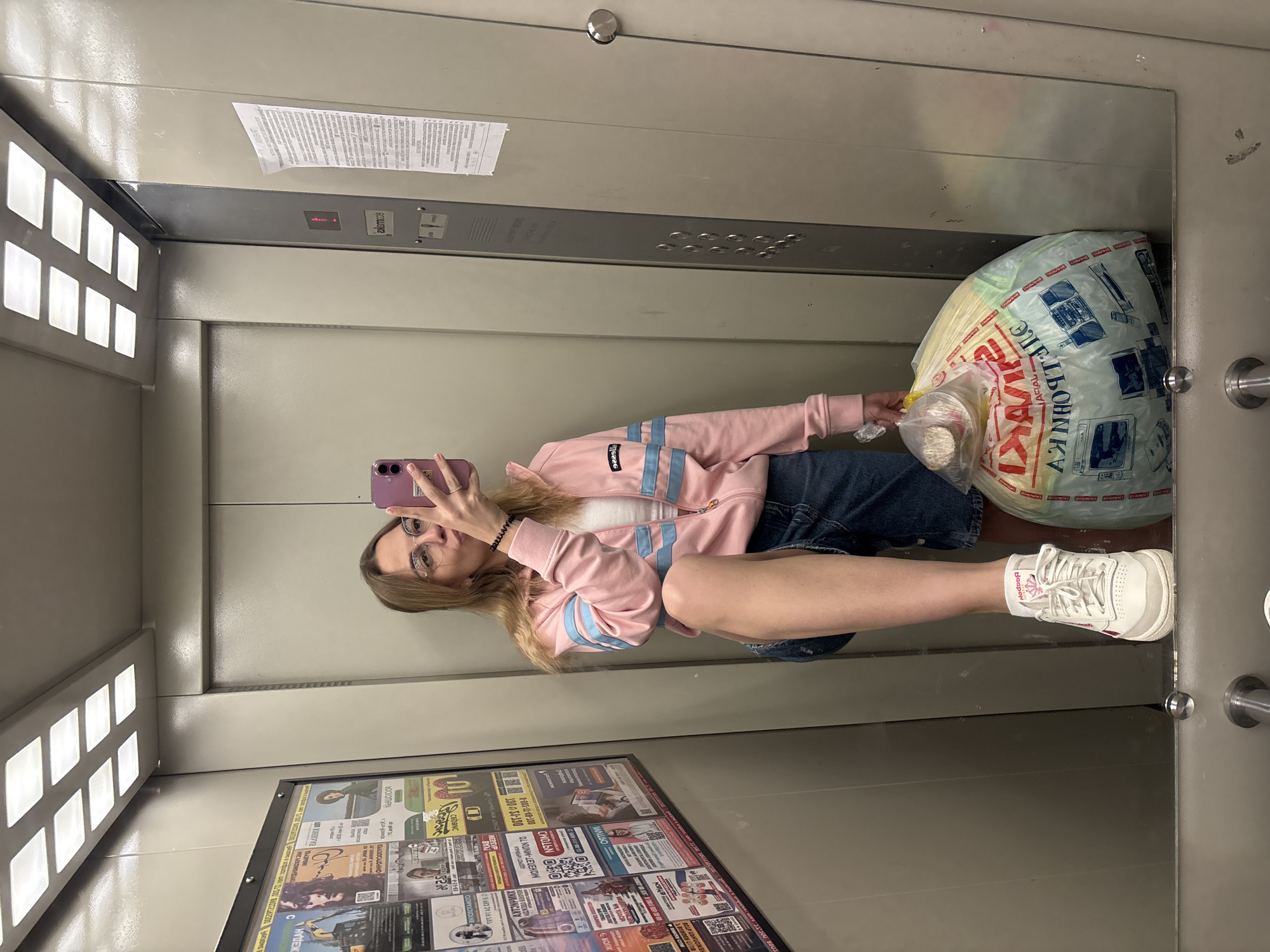 A young woman is taking a selfie in an elevator. She is wearing a pink tracksuit jacket with light blue stripes, a white top, denim shorts, and white sneakers with pink accents. She is holding a purple smartphone in her left hand, pointing it towards the mirror. She is standing to the side, holding a large plastic bag filled with what appears to be a variety of items, including food or groceries. The elevator has advertisements on the lower portion of the wall on the left side, and a small panel with floor indicators is visible above. The walls are a light gray color.
Based on the Cyrillic script on the bag and advertisements, the location is likely in a Russian-speaking region. One of the advertisements contains Cyrillic text with what appears to be a phone number including the digits "067-04-17-300," which suggests Ukraine or a similar region as it is not a valid Russian code. A brand name "Reebok" appears on the shoe she is wearing.
The elevator appears to have a floor indicator showing "1".
Given the combination of elements from the OCR'd Cyrillic text, including "Ust-Ilimsk" (likely a place name in Russia) appearing in the advertisement, it is probable that the scene takes place in Ust-Ilimsk, Russia.