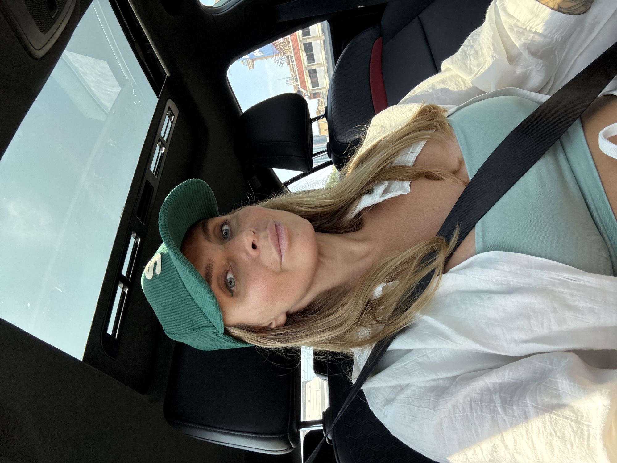 A woman wearing a green corduroy baseball cap with an embroidered "S" and a light blue tank top is in the driver's seat of a car. She has on an unbuttoned white linen shirt over her tank top. Her seatbelt is fastened across her chest. The car has a sunroof, which is open, allowing sunlight to illuminate the interior. Outside the car windows, buildings and a tall structure, possibly a hotel or office building with a sign that reads "OO," are visible, suggesting an urban environment in Bangkok, Thailand. The interior of the car appears to be leather with red accents on the seats.