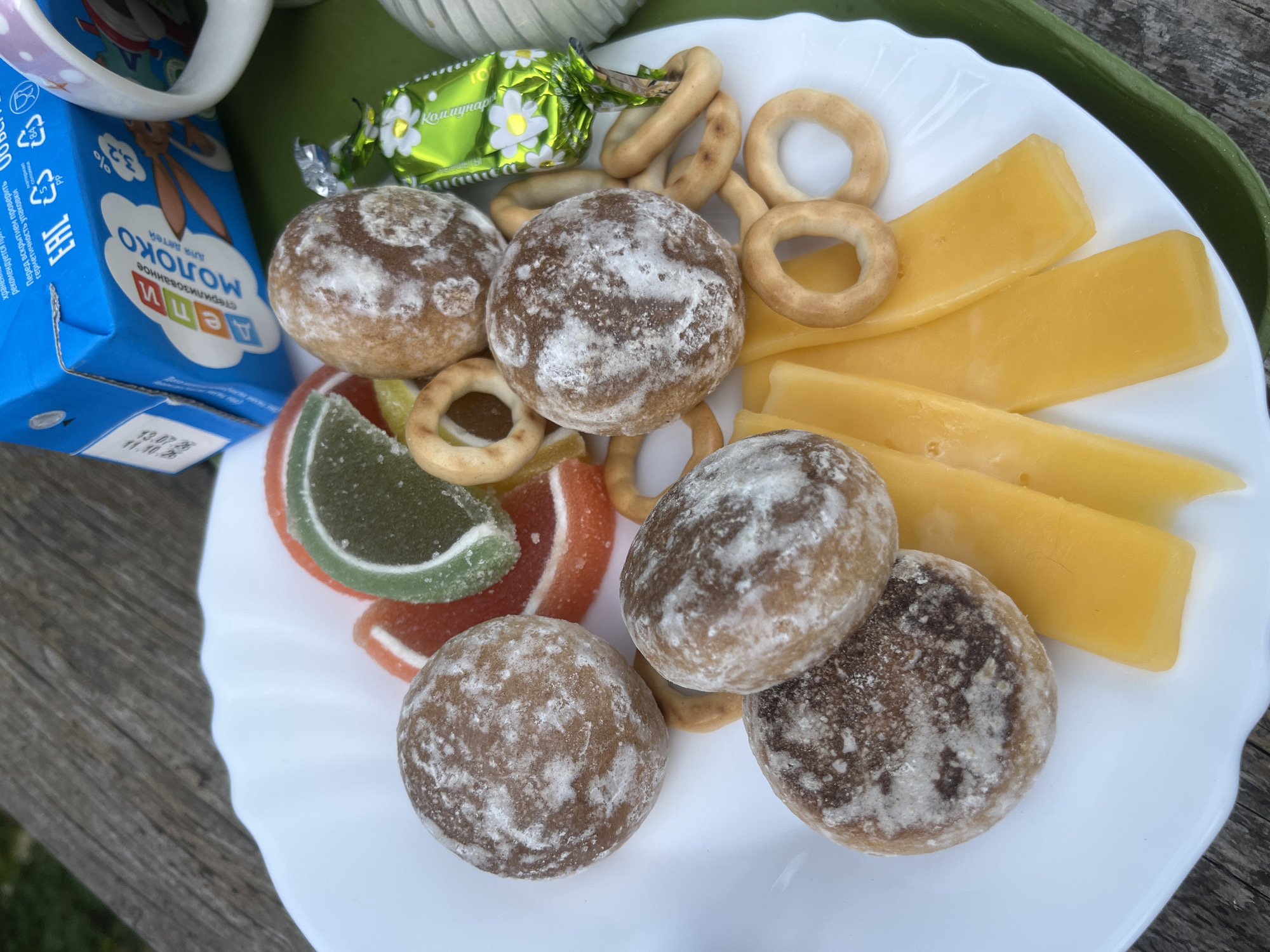 On a white plate, which is situated on a green tray placed on a wooden surface, a collection of snacks is arranged. The snacks include several round, brown cakes dusted with white powder, some pieces of orange and green jelly, small ring-shaped crackers, and slices of yellow cheese. A green candy wrapper with a floral pattern is also visible. In the background, a blue carton of milk labeled "Молоко для детей" (Milk for children) and part of a white mug are present. The setting appears to be outdoors, possibly a picnic or an outdoor meal. The overall scene depicts a light meal or snack spread.