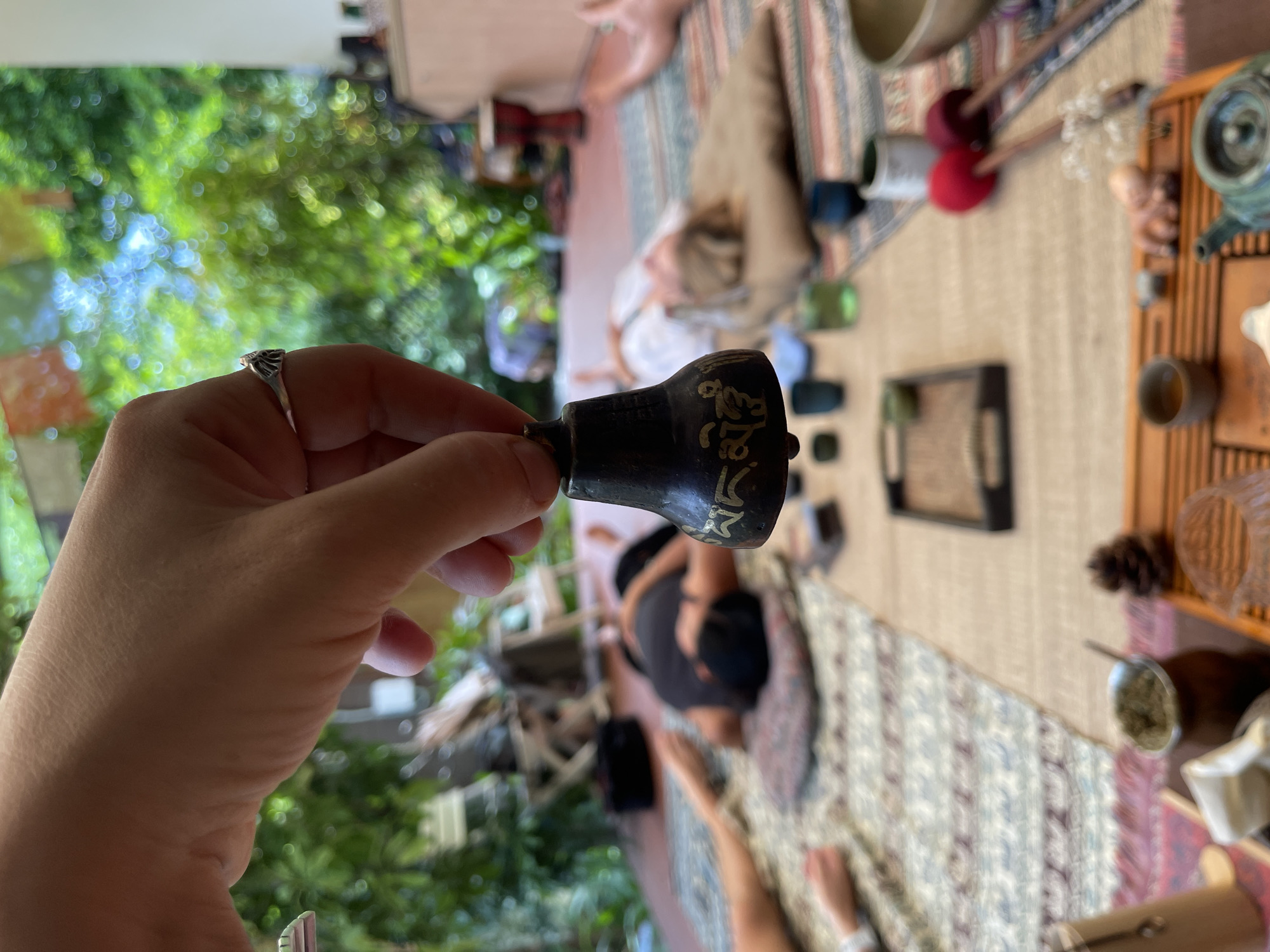 A first-person perspective captures a hand holding a small, dark bell adorned with ornate golden writing, possibly a mantra. The hand wears a silver ring on its index finger. In the blurred background, several people are visible, lying or sitting on patterned mats laid out on a wooden floor. Some appear to be participating in a meditation or yoga session, given the relaxed postures and serene atmosphere. The scene is illuminated by natural light filtering through lush green foliage, suggesting an outdoor or semi-outdoor setting.
The backdrop hints at a tranquil environment, possibly a garden or a patio area in Đà Nẵng, Vietnam. The presence of various objects like wooden trays, cups, and what appear to be singing bowls further reinforces the theme of a wellness or spiritual practice. The time of day seems to be daytime, with soft sunlight creating a warm ambiance. No specific weather conditions are apparent, but the overall mood is peaceful and contemplative. The golden script on the bell is illegible due to the image's resolution and the bell's texture.