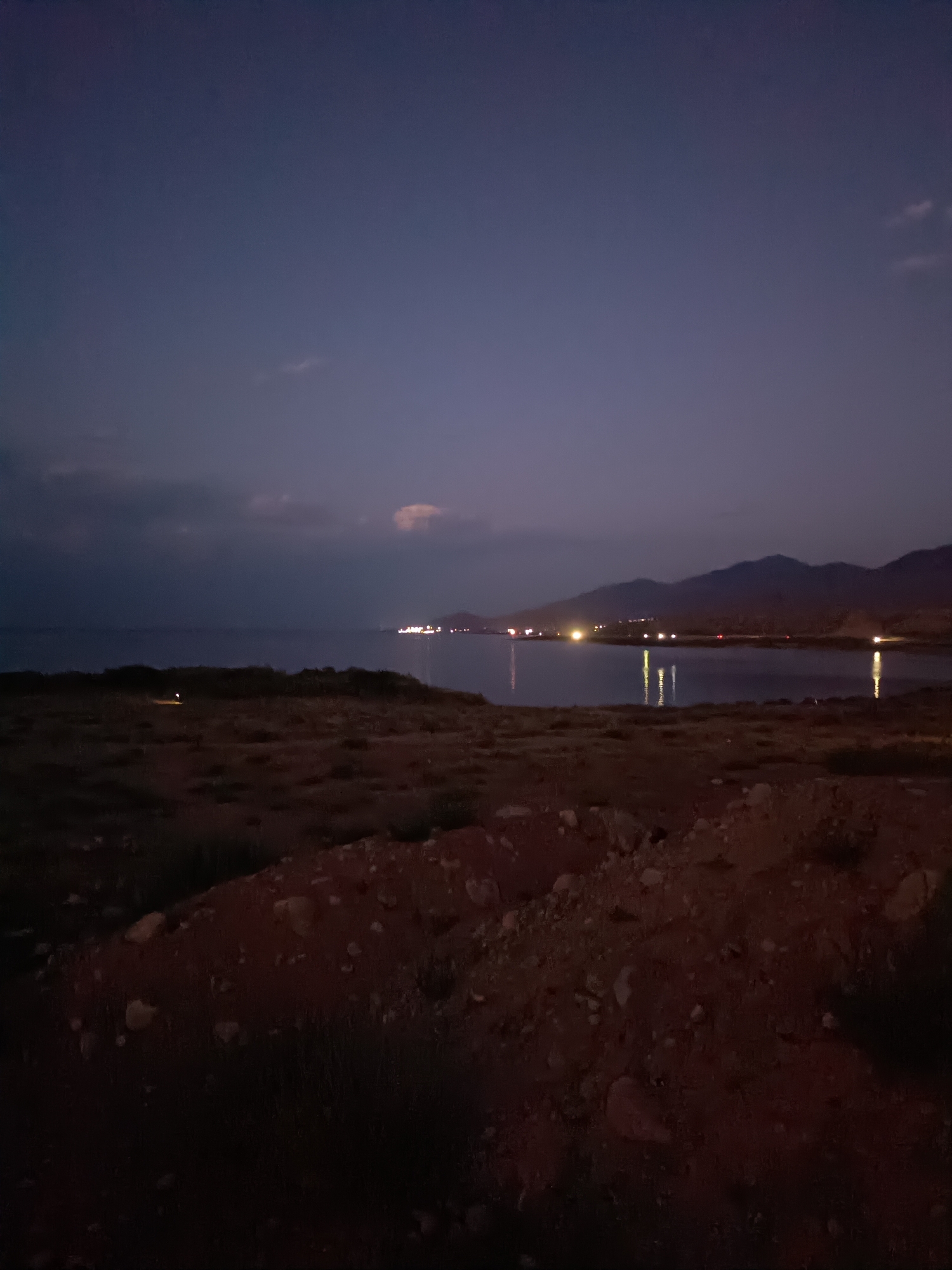 The image depicts a twilight scene over a body of water with a distant coastline. The sky is a deep indigo, transitioning to a lighter blue at the horizon. A few wispy clouds are visible. The water is calm, reflecting the lights from a settlement on the distant shore. These lights appear as bright yellow and white specks, some elongated due to reflection. The coastline is mountainous, with dark silhouettes of hills against the sky. In the foreground, the terrain is rough and rocky, with patches of sparse vegetation. The overall atmosphere is serene and tranquil. The location is identified as Tong, Kyrgyzstan, suggesting a natural landscape near the Issyk-Kul lake.