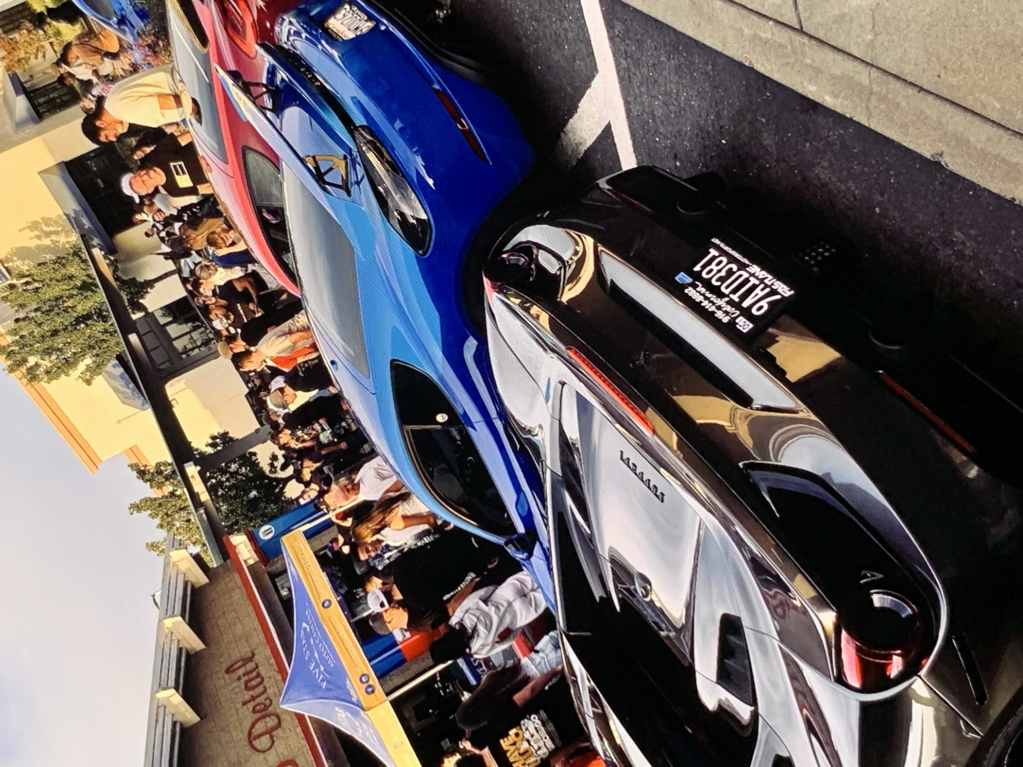 An outdoor gathering is underway in front of a building that appears to be a business with the word "Detail" visible on its facade. A blue sports car is prominently featured, parked alongside a black sports car in the foreground. Both vehicles are highly reflective. The black sports car has a license plate reading "9AID381 California" and a smaller plate that says "FASTLANE MOTORSPORTS.NET". Several people are congregated around the cars, with some appearing to be conversing and others observing the vehicles. A blue and yellow tent is set up to the left of the scene, with text on it that includes "FIVE STAR AUTO CENTER". The sky above is overcast, and trees are visible in the background, suggesting an urban or suburban setting during daylight hours. The scene appears to be a car show or a car enthusiast meetup.