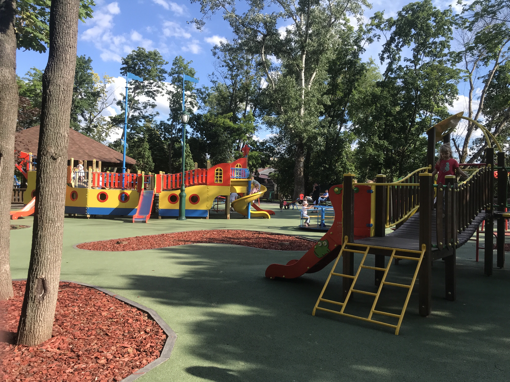 This is a full shot of an outdoor children's playground in Kharkiv, Ukraine. The playground has a large, colorful structure resembling a ship with yellow and red elements, multiple slides, and climbing areas. There are several children playing on the equipment, including a girl in a pink shirt on the right side of the image. The ground is covered in green rubber safety surfacing with areas of red wood chips. Tall trees with lush green foliage surround the playground, and the sky is blue with scattered white clouds. The overall scene depicts a lively and well-maintained recreational space.
