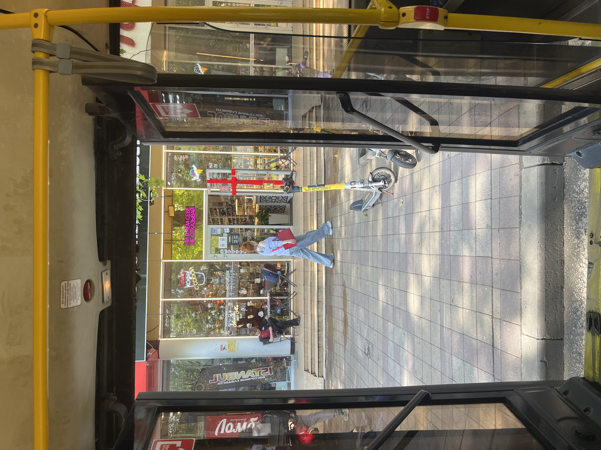 Here's a description of the image:
The scene is viewed from inside a bus, looking out through an open door onto a city street. The location is Krasnodar, Russia, based on the Cyrillic text visible in the storefront signs. A woman is walking across the sidewalk. Two electric scooters are parked or lying on the ground nearby, one branded "WHOOSH." There is a storefront visible with a red cross symbol above the door. The windows have displays with food items, and there are tables and chairs where customers are seated. Storefront signs are written in Russian. The bus's interior is visible, including yellow handrails and a stop button.