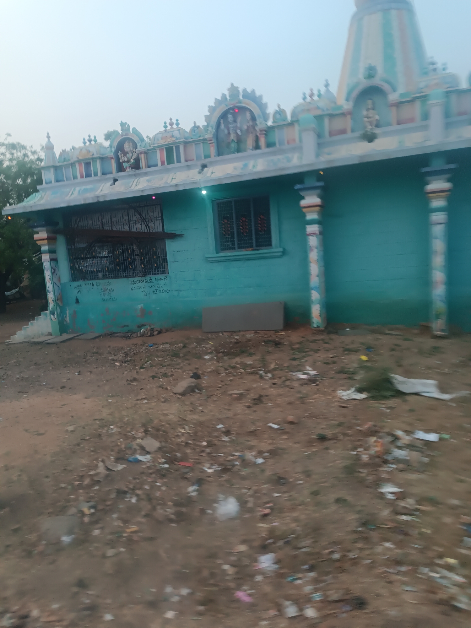 This image displays a teal-colored religious structure, likely a temple or shrine, situated in Kottalapalli, India. The building features a flat roof adorned with multiple colorful sculptures of Hindu deities positioned within arched niches, and a prominent, multi-tiered gopuram-like spire is visible on the right side of the roofline. The facade is painted a uniform teal/turquoise.
Two windows are present: a larger, rectangular window on the left with black metal security grates and a horizontal brown wooden beam, and a smaller, rectangular window on the right, also secured with black grates. The lower section of the wall to the left of the main entrance displays writing or graffiti in a local script, possibly Telugu. Two decorative pillars, painted with vertical stripes in white, red, and blue/green hues, support the roof overhang on the right side of the building. A set of concrete steps is visible on the far left, leading to an obscured entrance. Two small, bright lights are affixed to the facade, one above the left window (emitting a pink/purple glow) and one closer to the right (emitting a green glow). A rectangular, grey panel leans against the lower wall of the building to the right of the left window.
The foreground consists of unpaved, uneven reddish-brown earth heavily littered with various types of refuse, including plastic bags, paper, wrappers, and discarded debris, with sparse patches of green vegetation. The sky above is a uniform, hazy light grey, suggesting an overcast day or atmospheric conditions such as mist or smog. A tree with green foliage is partially visible on the far left, behind the structure. No individuals or specific actions are discernible within the frame.