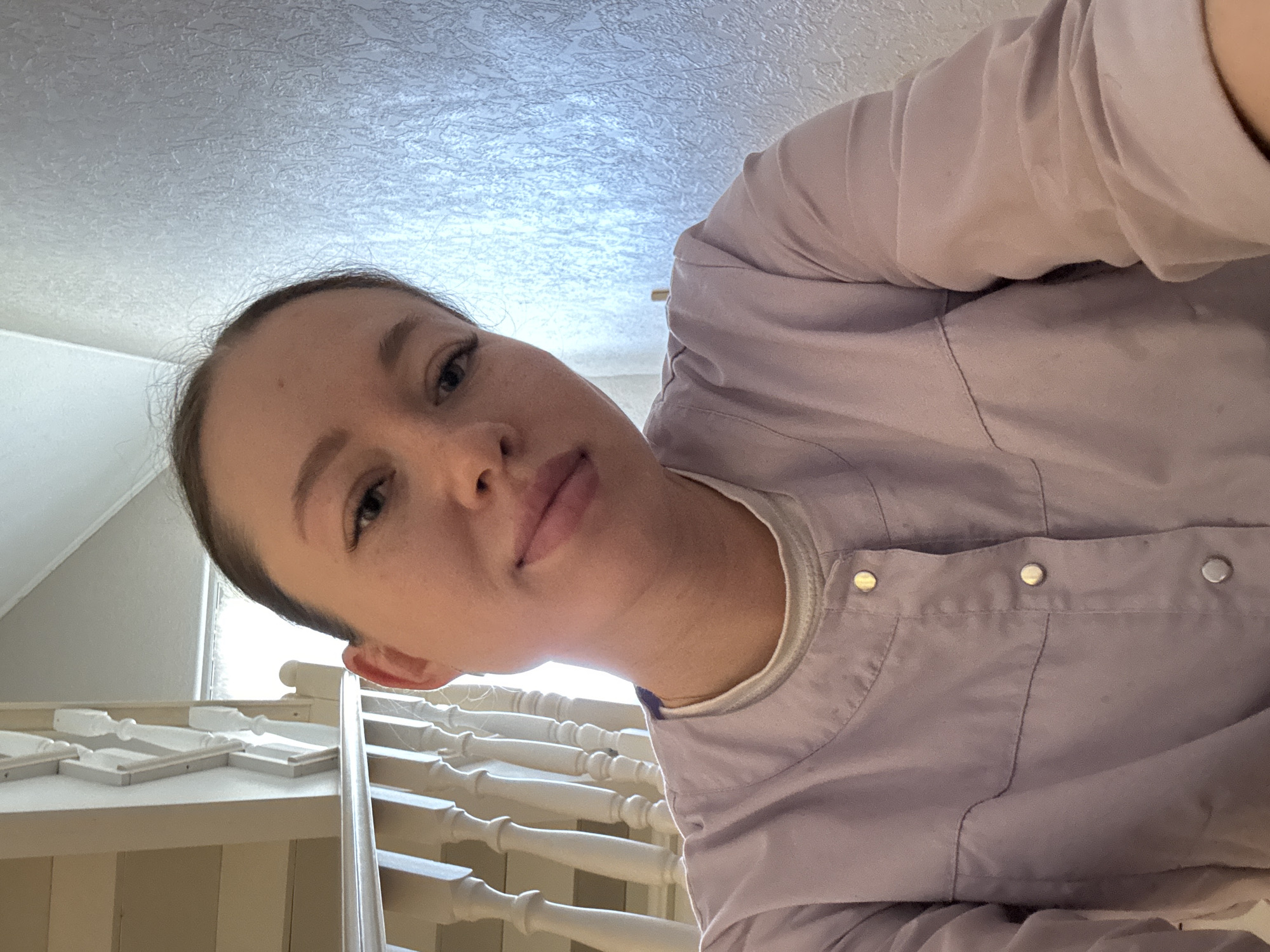 A woman is positioned above and looking down at the camera from a high angle, with a background that includes an interior residential space. The woman has fair skin, light brown hair pulled back from her face, and is wearing a light purple button-up garment over a white undershirt. Below her is a white staircase with decorative spindles and a railing. Above her, the ceiling is visible, showing a textured surface with a pattern resembling a light-colored material. A glimpse of an interior room or space can be seen in the upper left background. There is no definitive information to conclusively place the location specifically in Ферма, Russia.