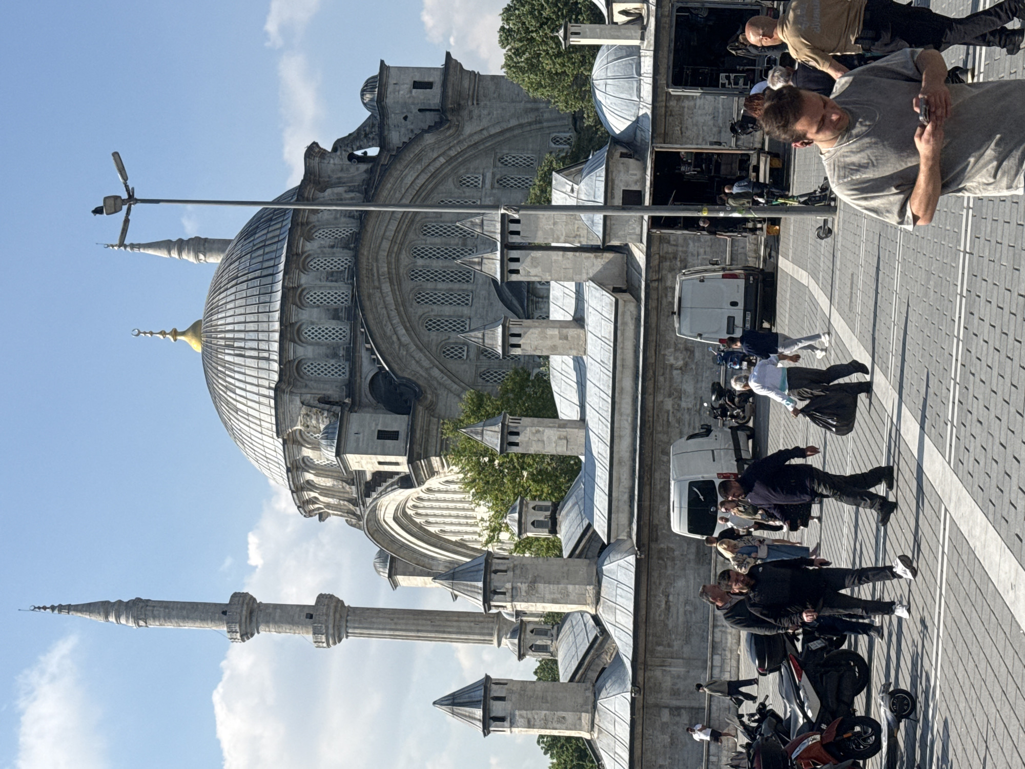 Here is a detailed description of the image:
The image presents a scene in what appears to be a public square or plaza in Unknown, Unknown, adjacent to a large, ornate mosque. The mosque features a large central dome, several smaller domes, and minarets, all typical of Islamic architectural design. The architecture suggests a historical and cultural significance of the location.
In the foreground and adjacent to the mosque, there's a paved area, possibly a pedestrian zone, where several people are walking or standing. There are some parked motorcycles and a white van. Several people are interacting with their smartphones. The presence of people and vehicles suggests an active public space.
The scene has a clear daytime setting with blue skies and some clouds, indicating good weather conditions.