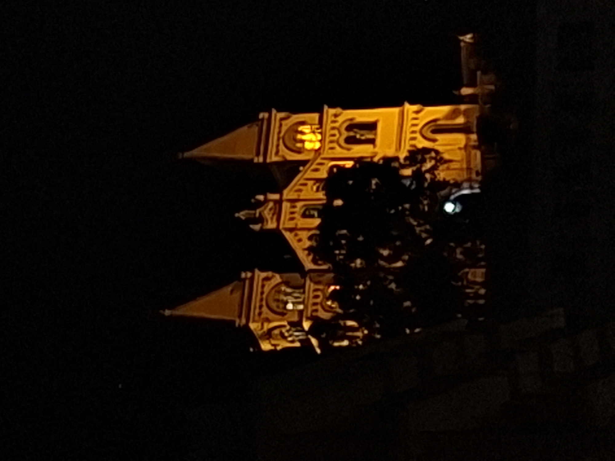 The image depicts a building, likely a church or cathedral, illuminated at night. The building features a spire, windows, and intricate architectural details highlighted by the lighting. A tree obscures a portion of the facade. The surrounding environment is dark, suggesting the image was taken in the evening. The location is Unknown, Unknown.