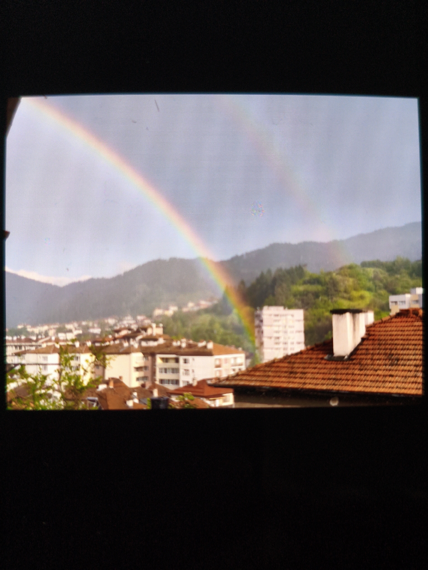 The image depicts a view from what appears to be a window, showing a cityscape with mountains in the background and a double rainbow in the sky. The rooftops of buildings are visible in the foreground, with a mix of residential and possibly commercial structures. The mountains in the distance are covered with trees. The sky is overcast. Based on the prompt, the location is Devin, Bulgaria.