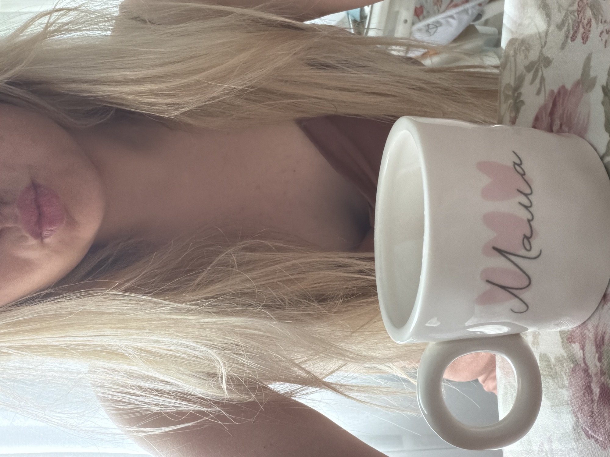 A light-skinned person with blonde hair partially obscures the view, with only their lips and part of their neckline visible. A white ceramic mug with pink heart decorations and the word "Mama" written on it is positioned to the right of the person. The mug sits on a floral-patterned surface. The location is not discernible from the image; there is no basis to determine the location as Votkinsk, Russia.