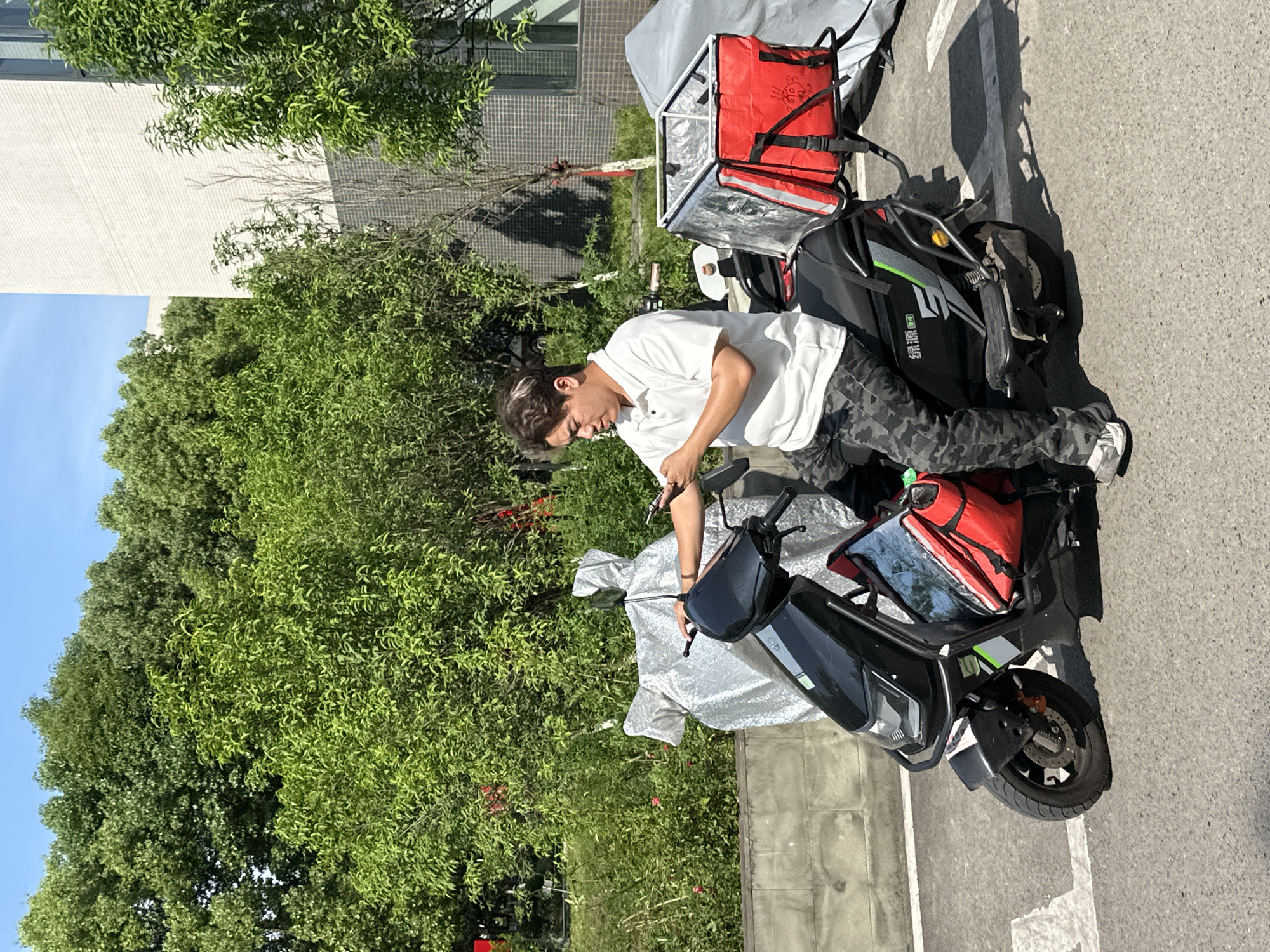 A young man is depicted standing outdoors next to a black electric scooter, bathed in bright sunlight. He is positioned on the right side of the scooter, leaning over the handlebars, seemingly engaged in an adjustment or preparation. He wears a white short-sleeved polo shirt, camouflage cargo pants, and white sneakers. His dark hair has lighter streaks and is styled casually.
The black electric scooter features prominent red delivery bags or containers attached to its rear, indicating it's likely used for delivery services. One bag is clearly visible, while another appears partially covered by a silver, reflective material draped over parts of the scooter. The scooter has green and grey graphic elements on its side, with the Chinese text "绿源" (Lü Yuan), a recognizable electric vehicle brand, clearly visible.
The scene takes place on an asphalt surface, possibly a street or parking area, with faint white markings. Strong, defined shadows are cast by the scooter and the man, suggesting a clear, sunny day, likely late morning or early afternoon. In the background, lush, dense green trees occupy a significant portion of the frame, hinting at a verdant environment. A light-colored building with a textured facade is partially visible behind the foliage. The sky is clear blue at the top left. The overall setting is consistent with an urban or suburban area in Jiangjin, China.