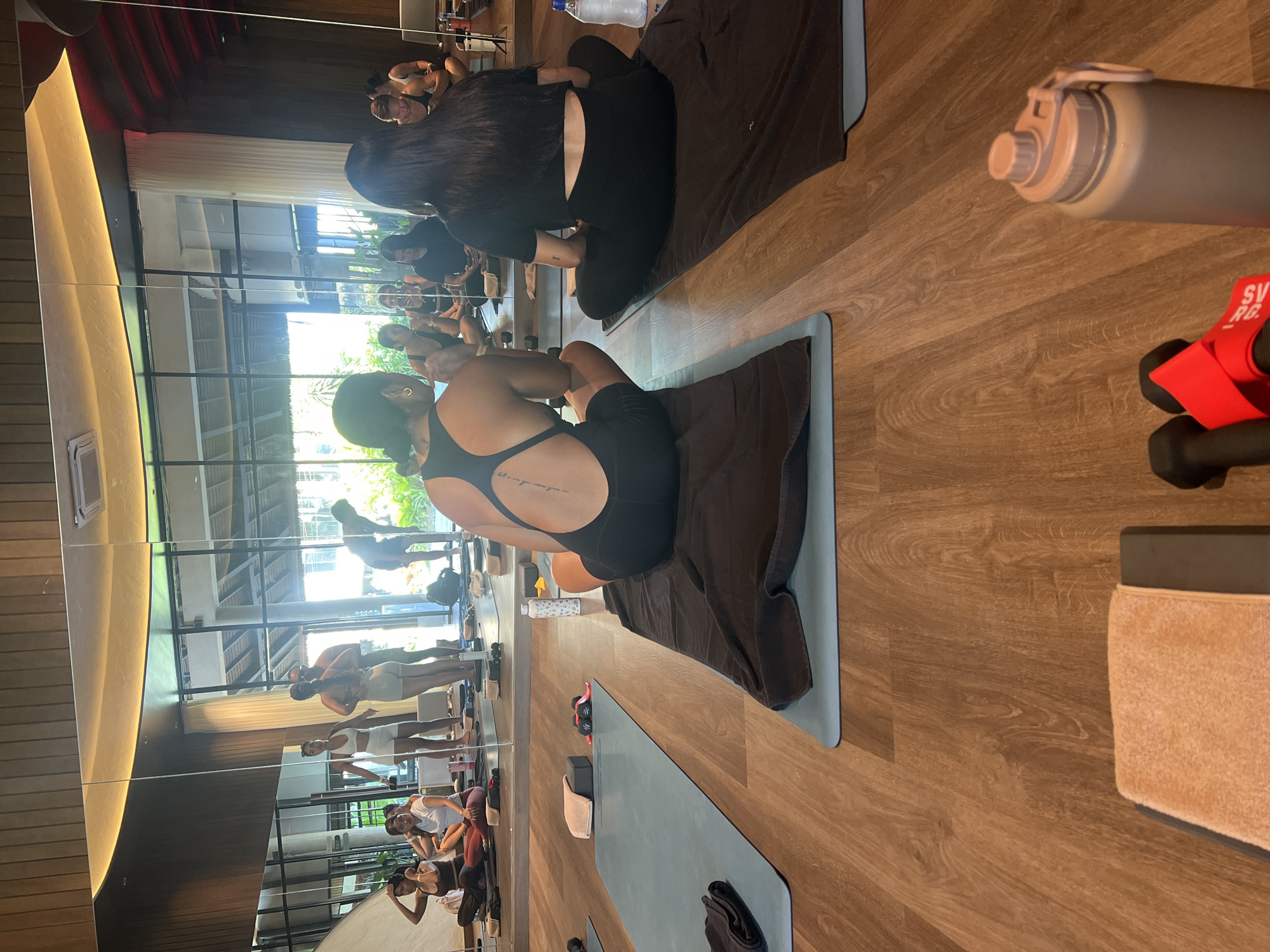 The image depicts an indoor yoga studio in North Kuta, Indonesia. The room features a wooden floor and a large mirrored wall that reflects the interior and the exterior through large windows. Several individuals are present, some participating in a yoga session and others observing. In the foreground, two women are seated on yoga mats, facing away from the camera. The woman on the left wears a black workout set with an intricate design on her back. The woman on the right wears a black crop top and leggings. Scattered around the room are yoga mats, towels, water bottles, and weights. The lighting suggests it is daytime, with natural light filtering in from the windows. The mirrors create a sense of space and amplify the reflections of the participants and the environment.
