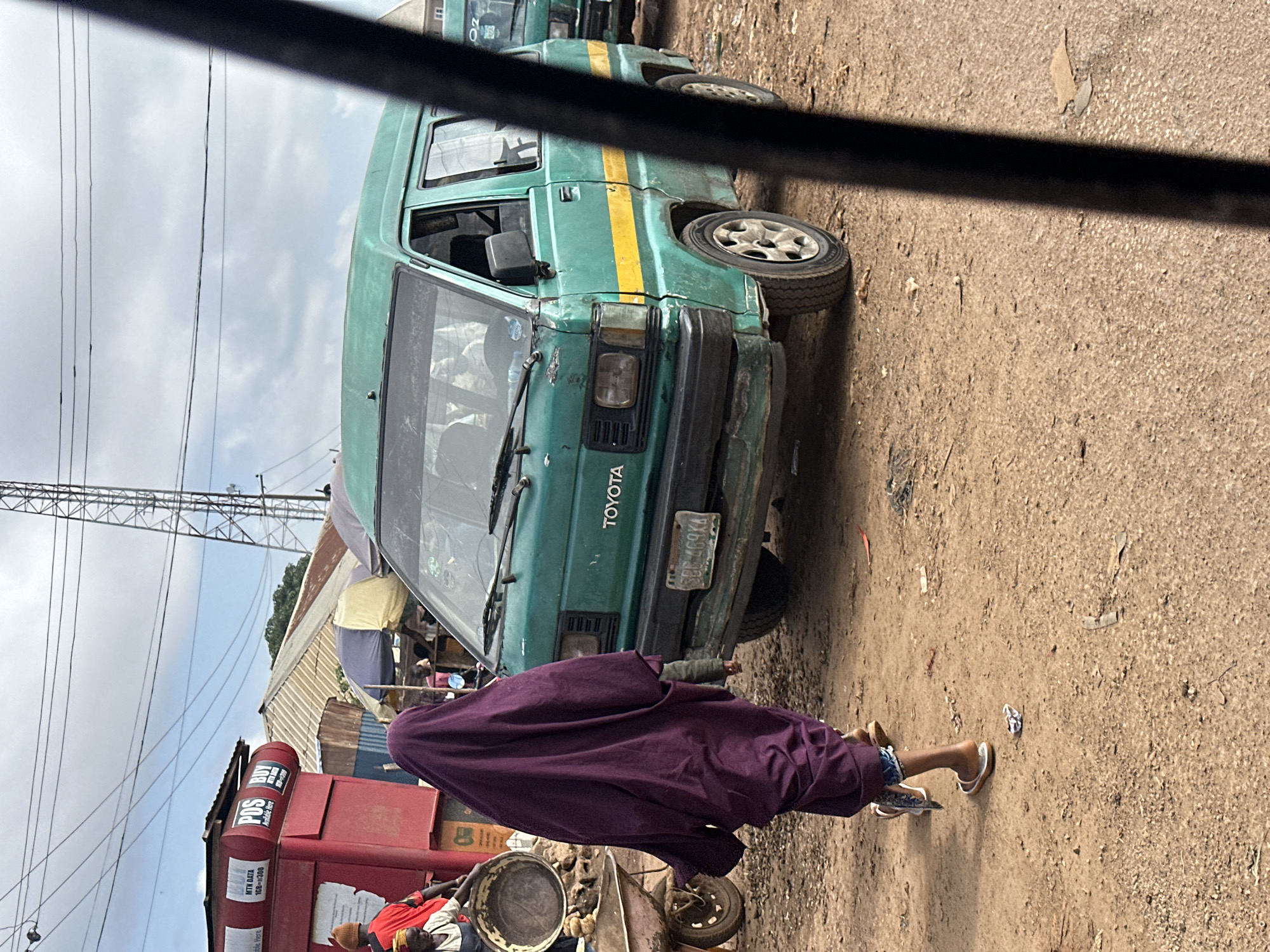This image captures a street scene in Jos, Nigeria, likely in a bustling market or transportation hub. In the foreground, a person wearing a dark purple hijab walks away from the camera, their feet clad in white flip-flops. To their right, a green Toyota Hiace van, common for public transport in Nigeria, is parked. The van has a yellow stripe running along its side and a license plate visible on the front bumper. Behind the van, another similar green van is partially visible. To the left of the frame, a red kiosk displaying "MTN DATA" and "POS Available Here" suggests commercial activity. The ground is unpaved and dusty, with scattered debris and a wheelbarrow nearby. In the background, several buildings with corrugated iron roofs and a telecommunications tower are visible under a cloudy sky. The overall impression is one of everyday life in a busy urban environment.