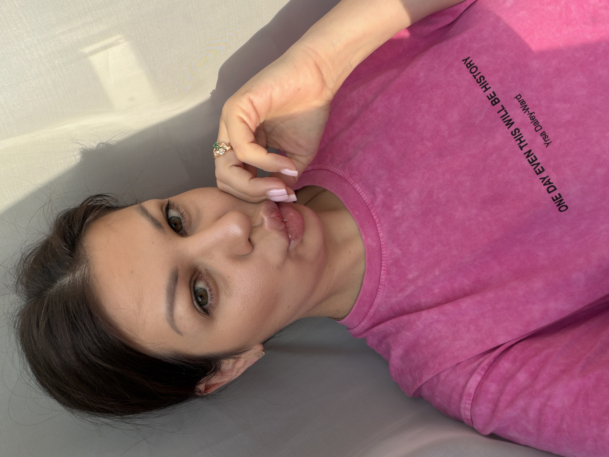 A selfie of a woman in Sochi, Russia, wearing a pink t-shirt with text on it. The woman is looking at the camera and puckering her lips. Her hair is dark brown and pulled back. She is wearing a ring with a green stone on her right hand. The background is a sheer curtain with shadows cast on it. The text on the t-shirt reads "ONE DAY EVEN THIS WILL BE HISTORY" and "Yrsa Daley-Ward".