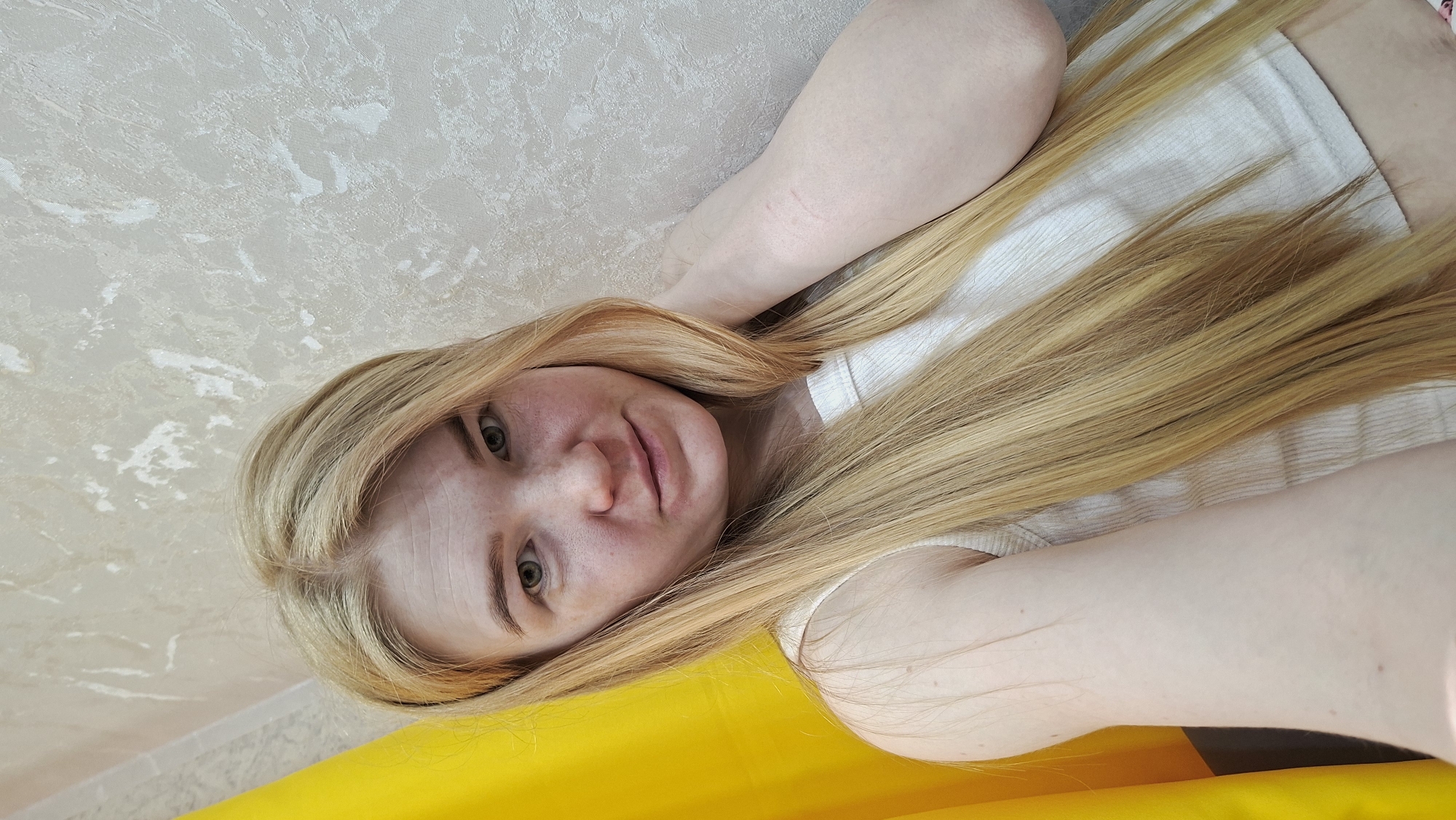 A fair-skinned woman with long blonde hair is lying down, taking a selfie-style photograph. She's wearing a light-colored tank top. The background includes a textured beige or cream-colored wall and a section of bright yellow fabric. The image gives the impression that the photograph was taken in a casual indoor setting, possibly within a residence. The picture appears to be taken at an angle. There is no indication of the location being in the city of Chaikovsky, Russia.