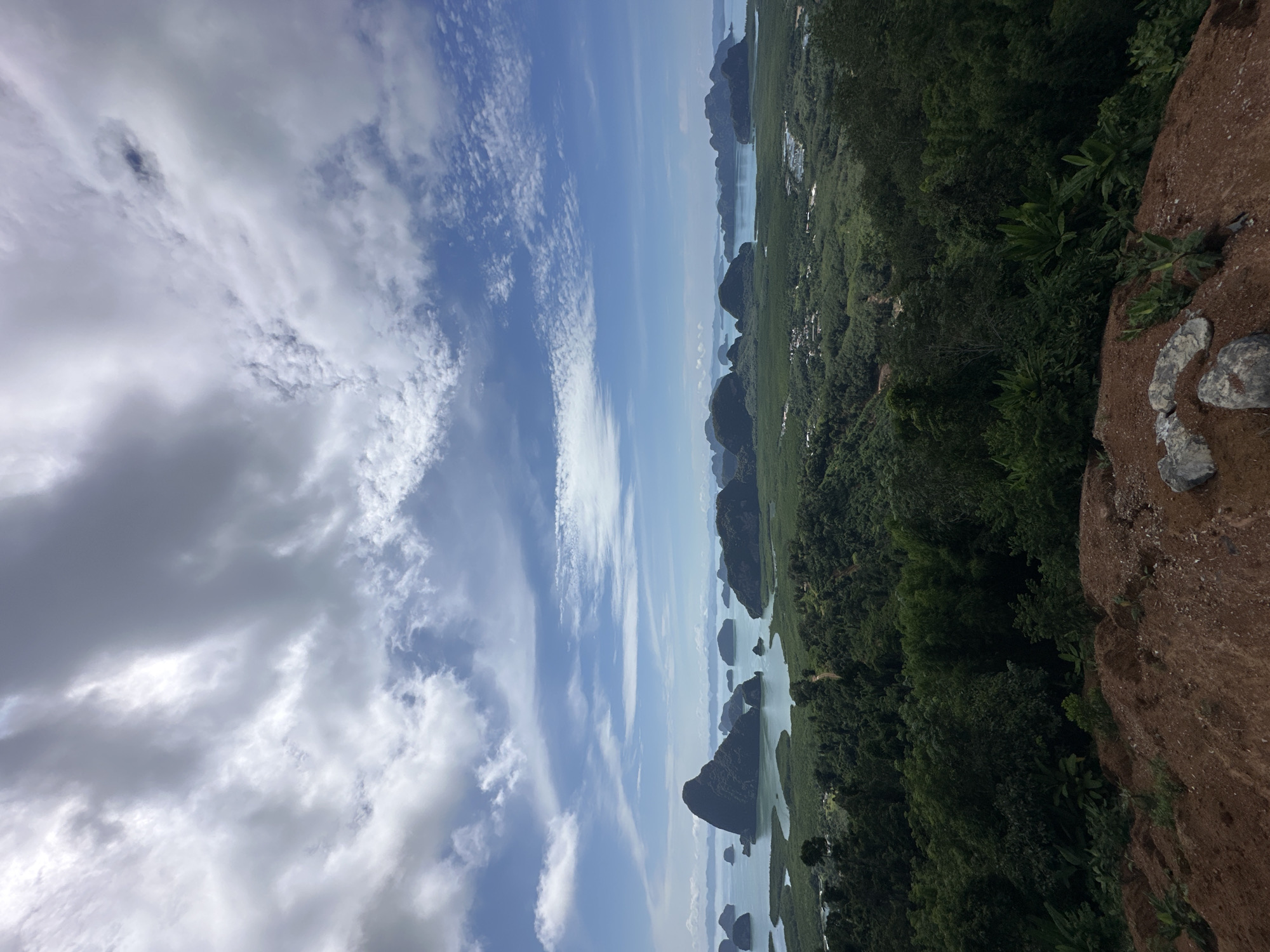 A high-angle, wide shot captures a scenic view of the Phang Nga Bay in Thailand. The foreground features a rough, brown dirt slope with scattered rocks and sparse vegetation. Beyond this, lush green trees densely cover rolling hills that descend towards the water. The bay itself is dotted with numerous limestone karst islands, some of which are quite large and prominent, with sheer cliffs rising from the turquoise water. The water appears calm and reflects the sky. In the distance, more islands and a hazy coastline are visible. The sky is a mix of blue sky and dramatic, white and grey clouds, suggesting a dynamic weather day. There are no people or animals visible in the frame. The scene conveys a sense of natural beauty and vastness.