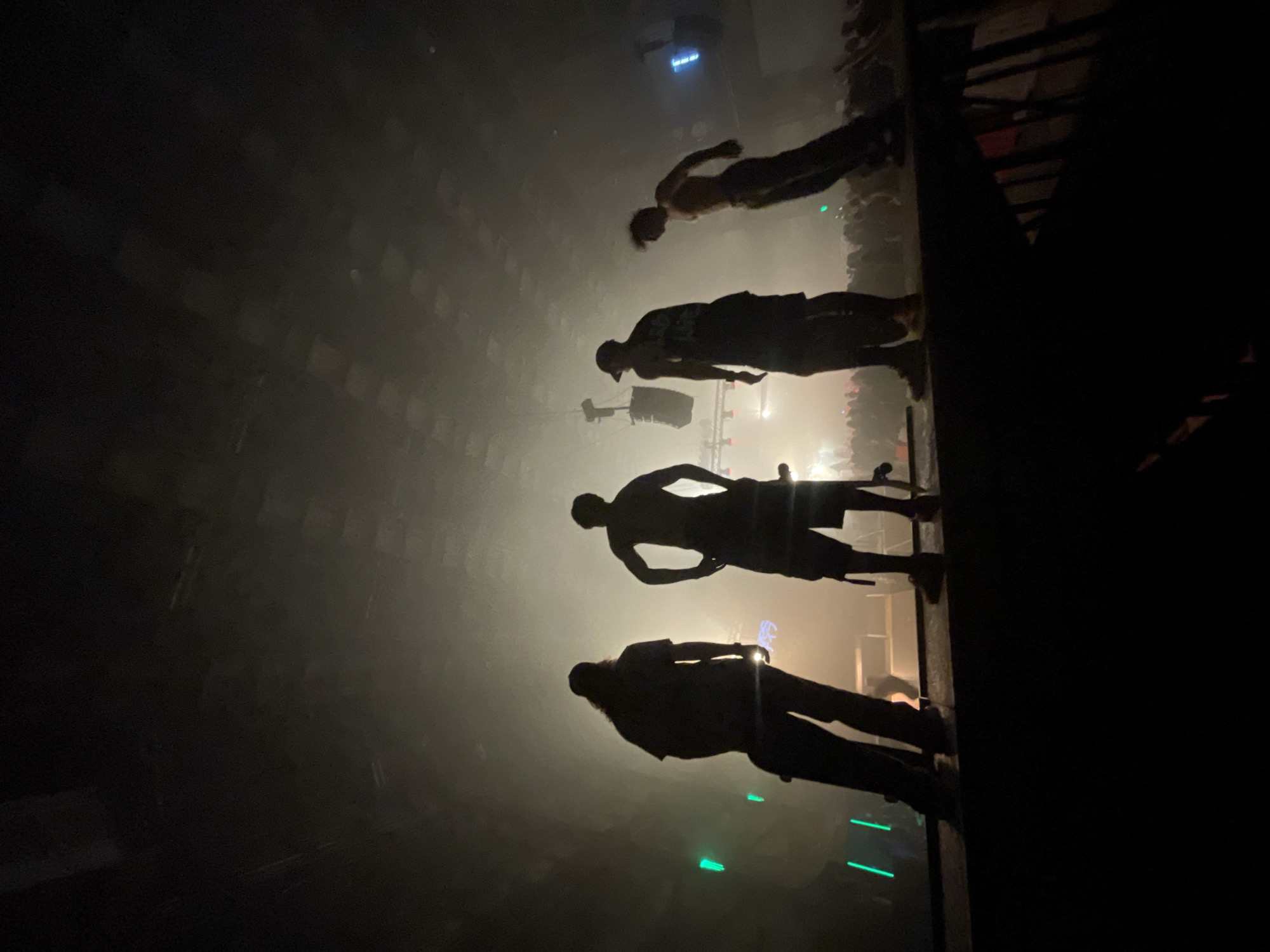 Four individuals stand silhouetted on a raised platform in a dimly lit, smoky venue, likely a concert or club. The lighting is atmospheric, with beams of light cutting through the haze and green lights visible on the left. The individuals appear to be men, two of whom are shirtless. One is holding what appears to be a skateboard. In the background, a crowd of people is visible, also in silhouette. The ceiling of the venue shows a checkered pattern of lights. There is no discernible indication of the city of Kyiv, Ukraine.