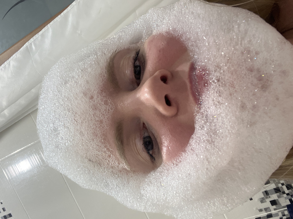 A close-up, low-angle shot presents a face surrounded by white, bubbly foam. The foam encapsulates the person's head, leaving the face visible. Eyes, nose, and lips are prominent features within the foamy enclosure. The background includes a white surface and a hint of white and blue tiled surface on the right side. The lighting is bright, causing the bubbles to glisten. The person appears to be relaxed.
