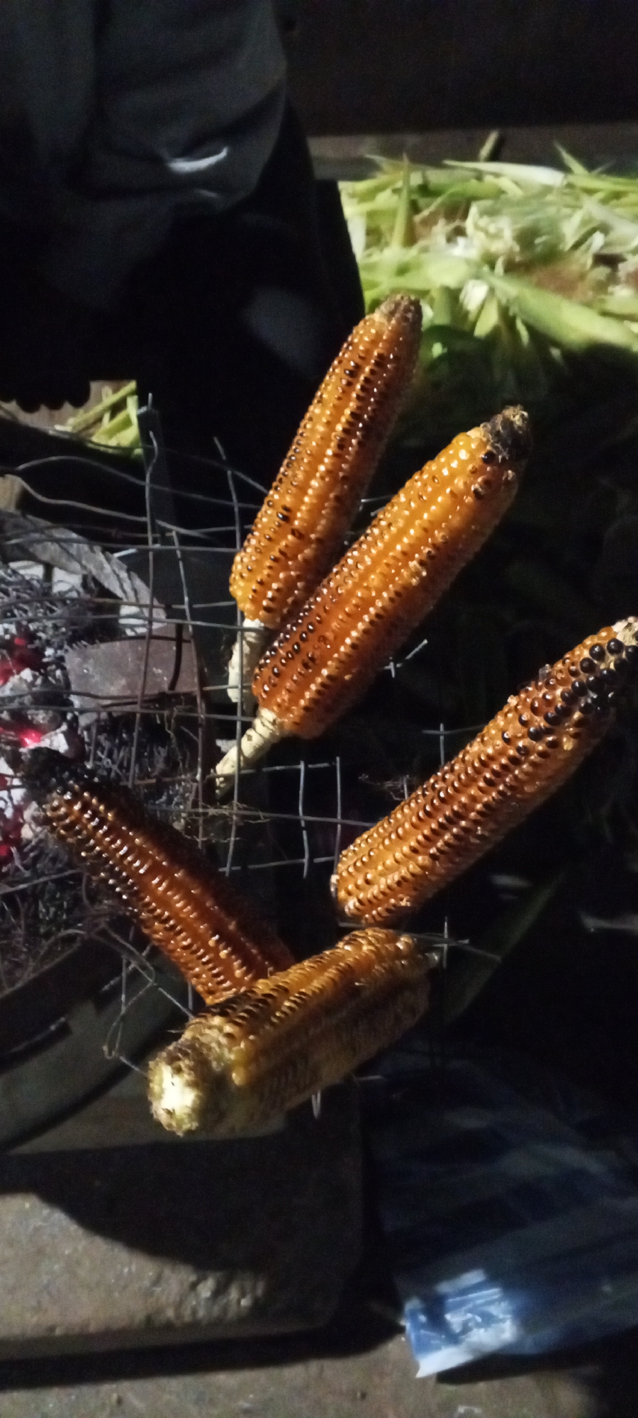A night shot shows several ears of corn being grilled over hot coals. The corn is arranged on a wire grill rack that is placed over a bed of glowing red coals. The corn cobs have a golden-brown color with some darker charred spots, indicating they are well-roasted. Some husks of corn are visible in the background, suggesting they were recently prepared for grilling. A person's dark clothing is partially visible in the upper left of the frame. The scene appears to be outdoors, possibly a backyard or a campsite. The overall impression is one of casual outdoor cooking.