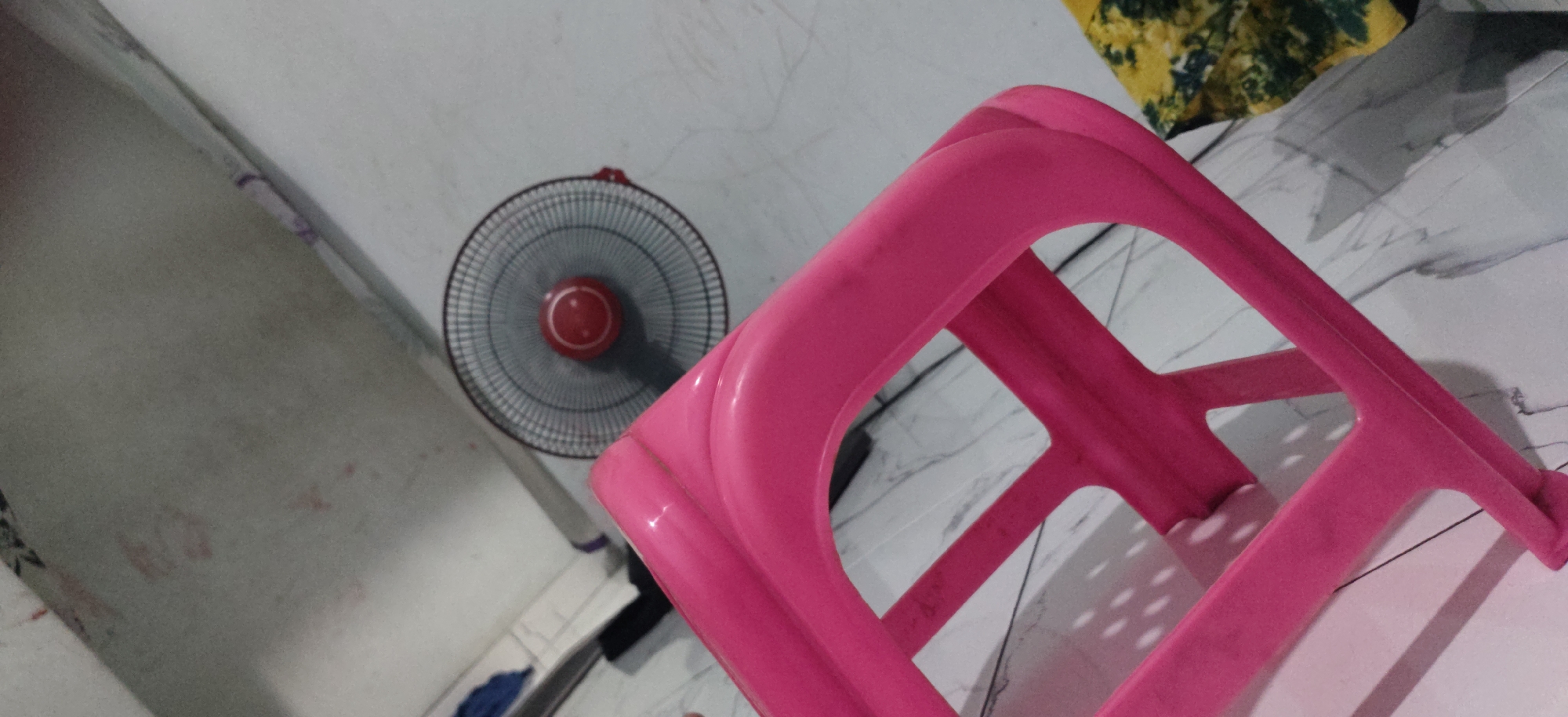 A bright pink plastic stool is positioned in the foreground, angled towards the right of the frame. The stool has a hollow, geometric design with interconnected bars. Behind the stool, on the left side of the image, a black oscillating fan with a red circular center is visible. The fan is angled slightly away from the viewer, and its blades are blurred, suggesting it may be in operation. The background consists of a pale, textured wall on the left, and a marbled-patterned floor on the right. A yellow and green patterned fabric is visible in the upper right corner, draped over an unseen object. The scene appears to be indoors, in a residential setting. No people are present in the image. The lighting is dim and creates shadows. The image was taken in West Jakarta, Indonesia.