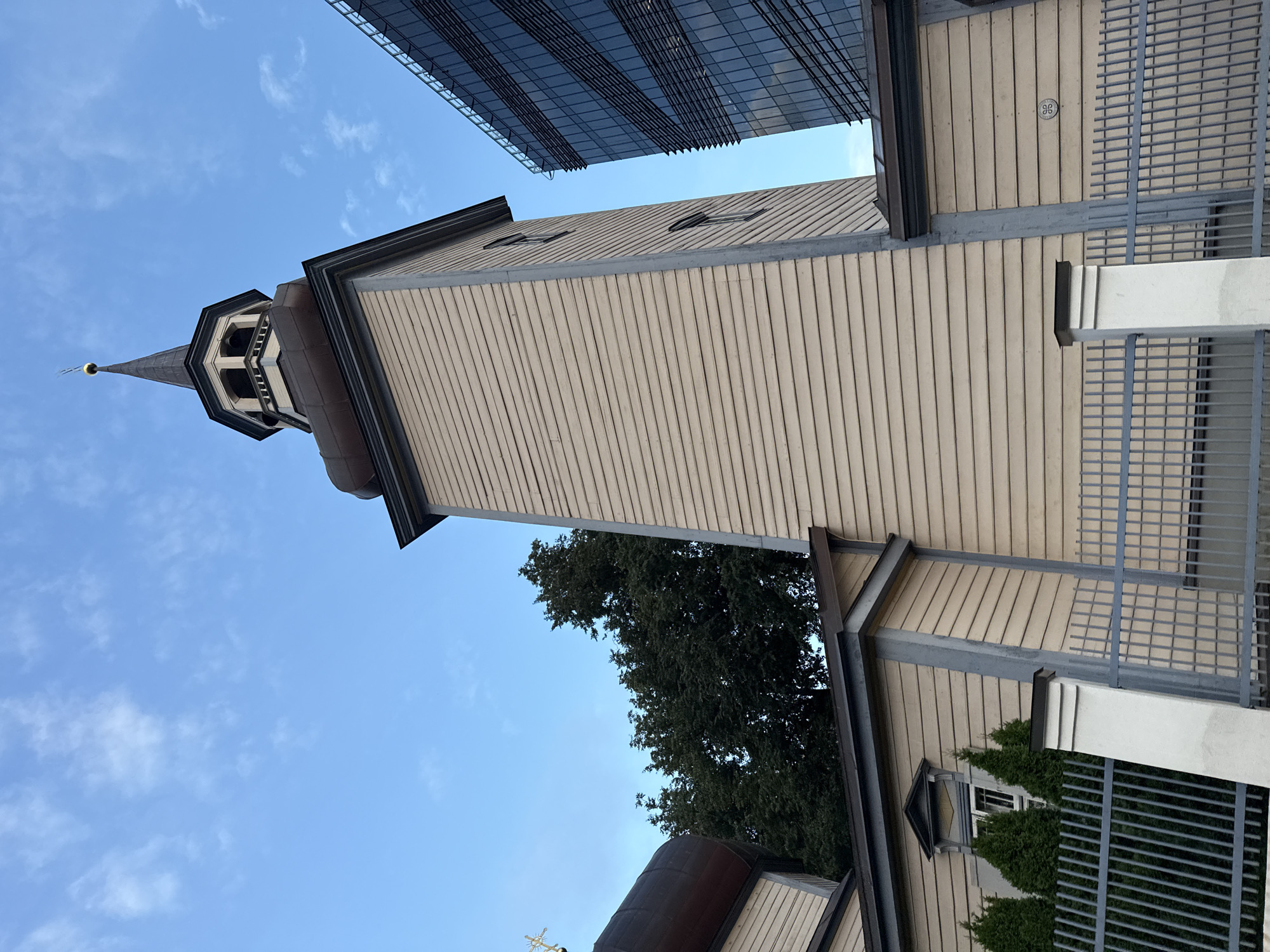 This is an outdoor, low-angle, eye-level shot of a wooden church tower and a modern glass building under a blue sky with scattered clouds. The church tower, painted a pale yellow with grey trim, has horizontal siding and a dark roof with a steeple topped with a golden cross. A bell is visible in an opening near the top of the tower. To the right of the church is a tall, modern building with a dark glass facade and horizontal metallic accents. A grey metal fence runs in front of the church, with a white pillar in the foreground. A few small evergreen trees are planted behind the fence. The scene appears to be in a city, with the contrast between the old church and the new building suggesting an urban environment. The location is likely in Tallinn, Estonia, given the architectural style and the known juxtaposition of historical and modern buildings in the city. There are no people visible in the image.