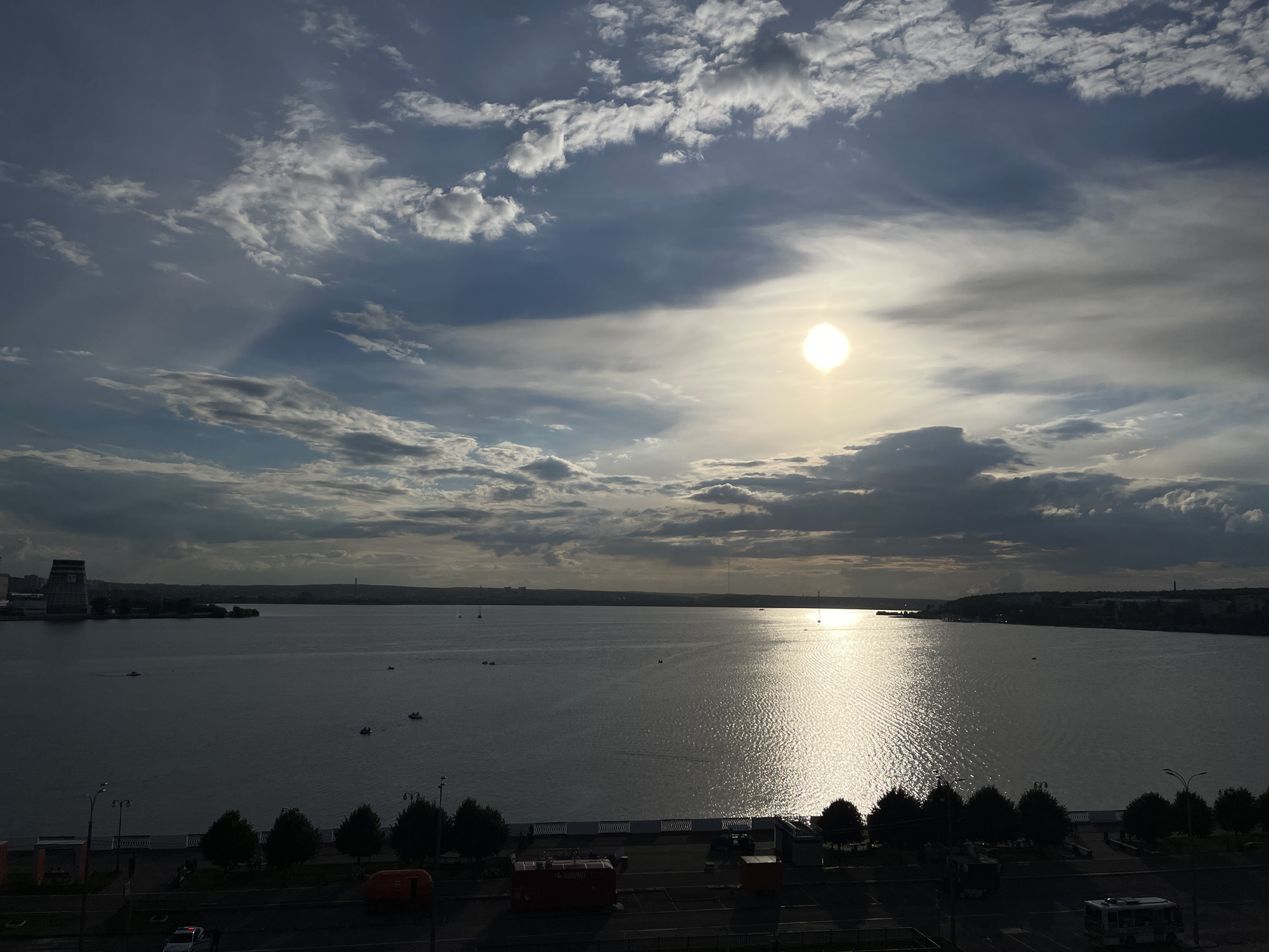 This is a high-angle, wide-angle photograph of a body of water, likely a river or lake, with a city skyline in the background. The sun is visible in the upper right quadrant of the sky, casting a bright reflection on the water. The sky is partly cloudy, with dramatic cloud formations. Several small boats are scattered on the water. In the foreground, there is a tree-lined promenade and a street with some vehicles and buildings. The overall impression is a city scene at sunset or sunrise, with natural beauty and urban elements coexisting.
