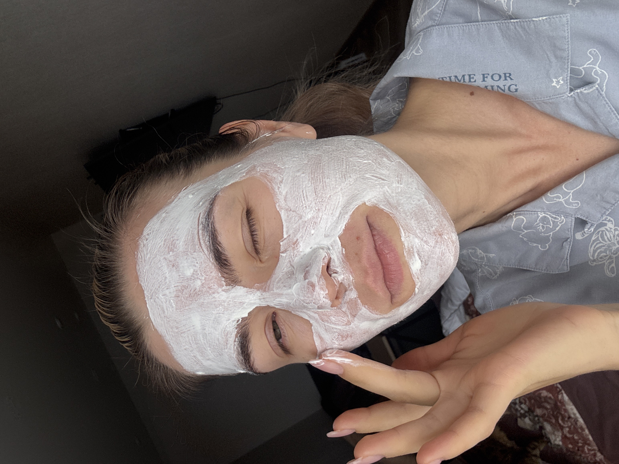 A fair-skinned person wearing a light blue pajama top with white cartoon bear designs and the words "TIME FOR RELAXING" is applying a thick white face mask. The mask covers the majority of the person's face, except for the lips and a small area around the nostrils. The person's eyes are partially closed. One hand, with manicured nails, is visible and has a finger touching the face mask. The background is dark and indistinct. The location and city are Unknown, Unknown.