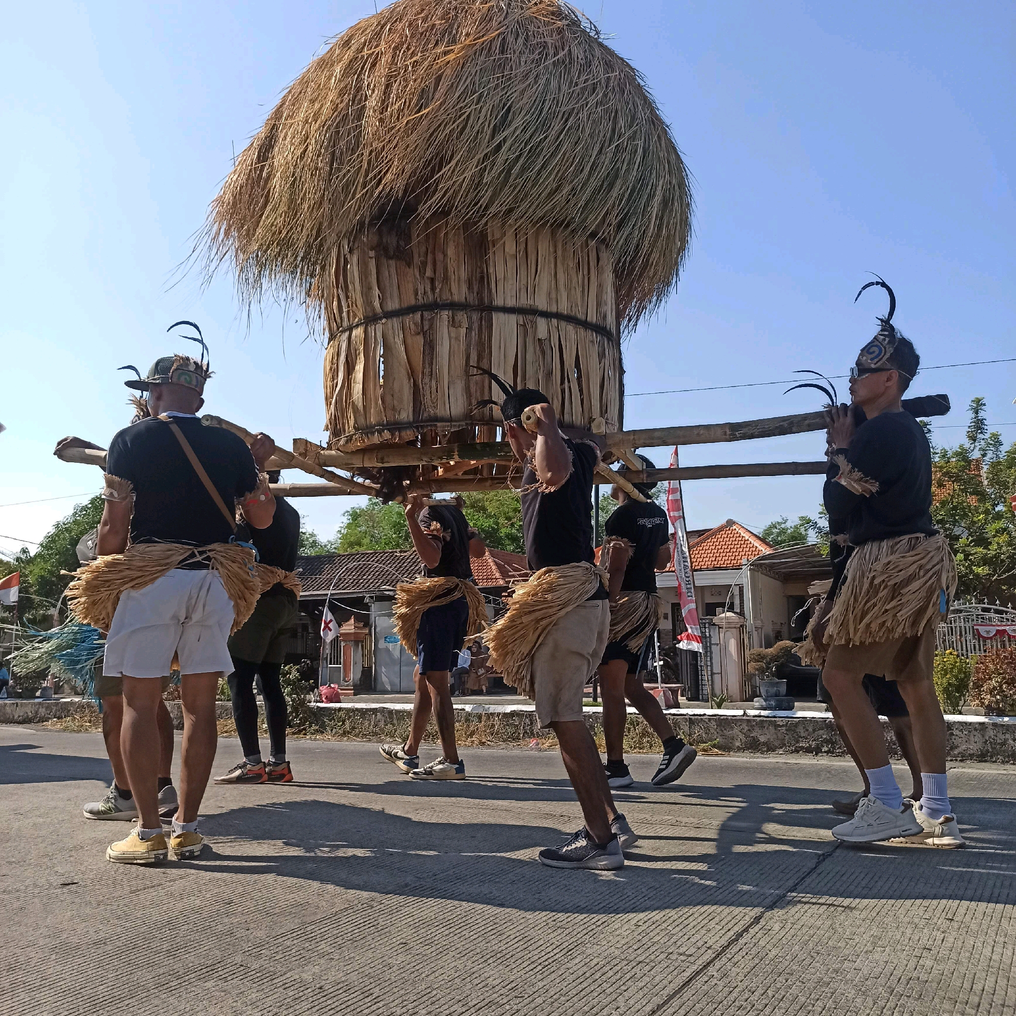 In Plaosan, Indonesia, a group of men dressed in traditional attire are participating in a parade. They are carrying a large, conical structure made of straw and dried palm leaves, supported by wooden beams. The men wear black t-shirts, white shorts, and straw skirts. Some of them have elaborate headdresses adorned with feathers. The parade is taking place on a paved road, with houses and trees visible in the background under a clear blue sky. Flags are also visible along the side of the road. The scene suggests a cultural or religious festival.