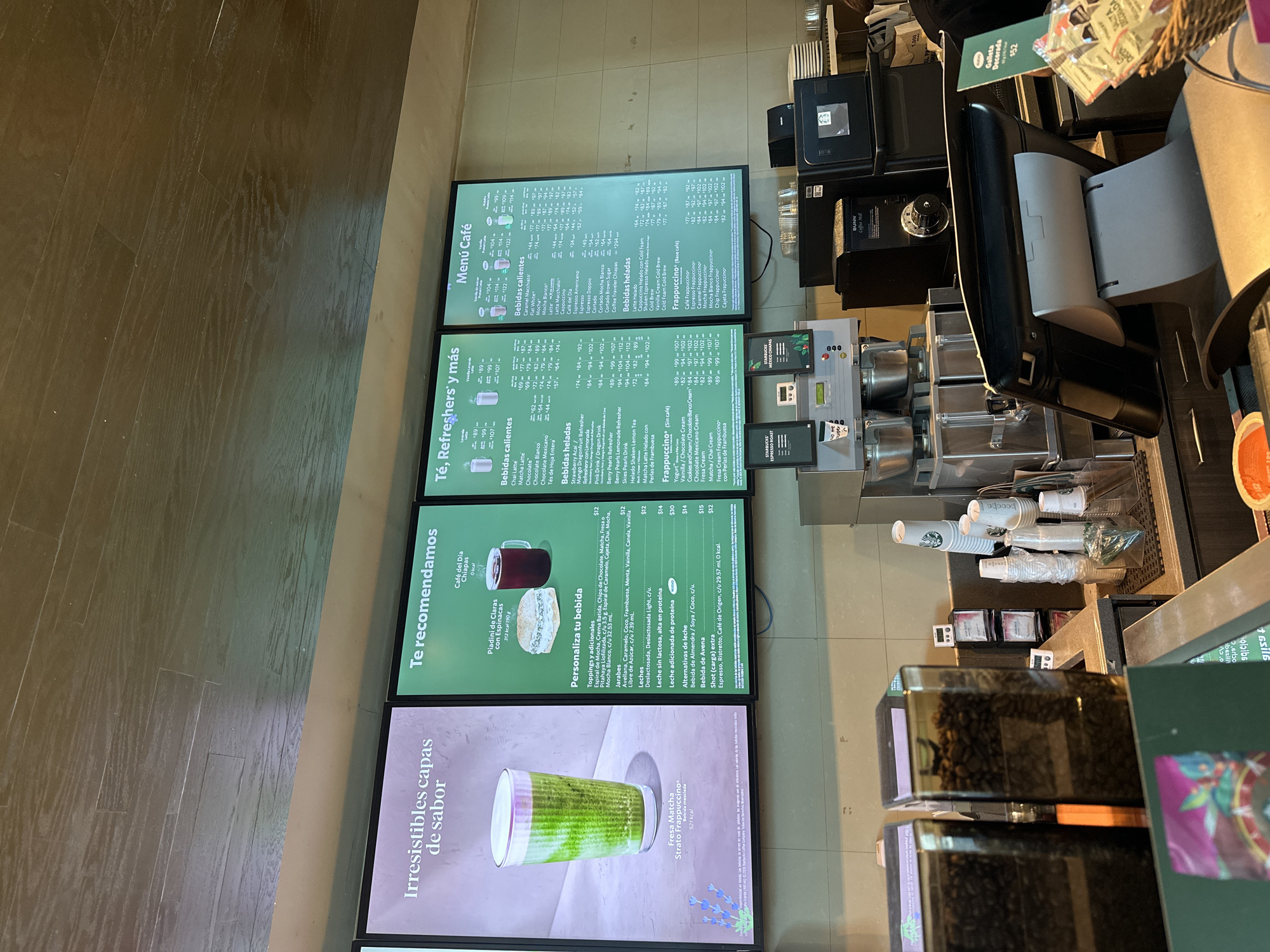 This media file captures the interior of a Starbucks coffee shop, specifically focusing on the menu displays and a portion of the counter area. No people are visible, but the setup implies a customer service and beverage preparation environment.
The scene is dominated by several vertically oriented digital menu boards. From top to bottom, these display "Menú Café" (Coffee Menu), "Té, Refreshers' y más" (Tea, Refreshers and more), "Te recomendamos" (We recommend), and "Irresistibles capas de sabor" (Irresistible layers of flavor), featuring a Fresa Matcha Strato Frappuccino. The menus list various hot and cold beverages, including coffees, teas, refreshers, and Frappuccinos, along with options to personalize drinks with toppings, syrups, and different types of milk (Leches, Alternativas de leche). Food items like "Piadini de Claras con Espinacas" (Egg white piadini with spinach) and "Café del Día Chiapas" (Coffee of the Day Chiapas) are also advertised. Prices are prominently displayed in pesos, ranging from single digits to hundreds (e.g., $12, $14, $30, and various price ranges for different sizes).
On the counter to the right, coffee-making equipment, including coffee grinders (one labeled "BUNN Coffee Mill") filled with coffee beans, a POS system, and stacked cups, are visible. A small basket containing sugar packets and stir sticks sits next to a display for a "Galleta Decorada" (Decorated Cookie) priced at $52. Further up, signs for "STARBUCKS ESPRESSO ROAST" and "STARBUCKS MÉXICO CHIAPAS" coffee are visible. The flooring appears to be a dark wooden or wood-patterned surface. The Spanish language on the menus, coupled with the "México Chiapas" coffee label and the provided location context of San Andrés Cholula, confirms this is a Starbucks in Mexico.