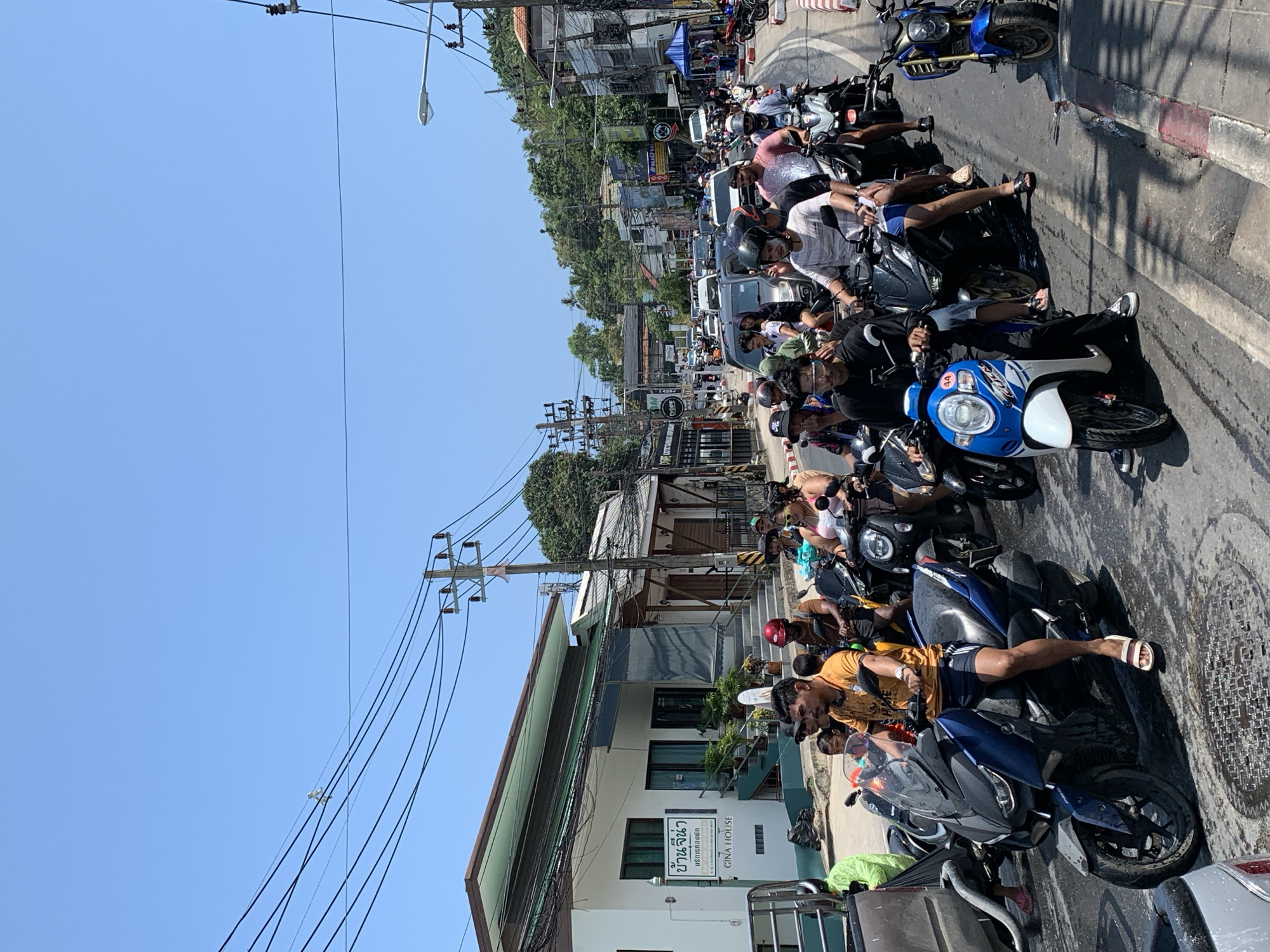This image shows a street scene in Baan Hua Thanon, Thailand. Multiple people are sitting on parked motorcycles lined up along the side of a street. Many individuals are wearing casual clothing, including shorts and t-shirts. The motorcycles are a variety of makes and models. Overhead power lines run along the side of the street. Buildings and trees are visible along the background. The sky is clear and blue. The light suggests it is daytime. The road surface is paved.