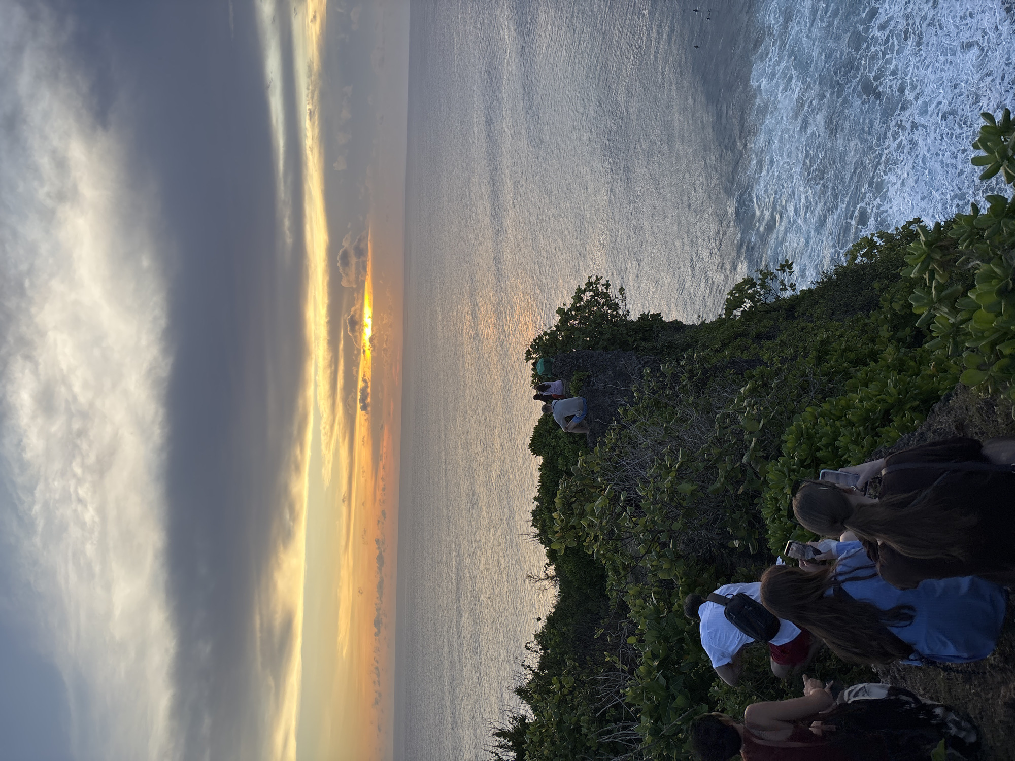 The photograph depicts a sunset scene at Uluwatu, Indonesia. A group of people are standing on a cliff overlooking the ocean. The sky is filled with clouds, and the sun is setting, casting a golden light across the water. Waves are crashing against the shore. Some individuals are capturing the scene with their phones. The vegetation is lush and green, covering the edge of the cliff.