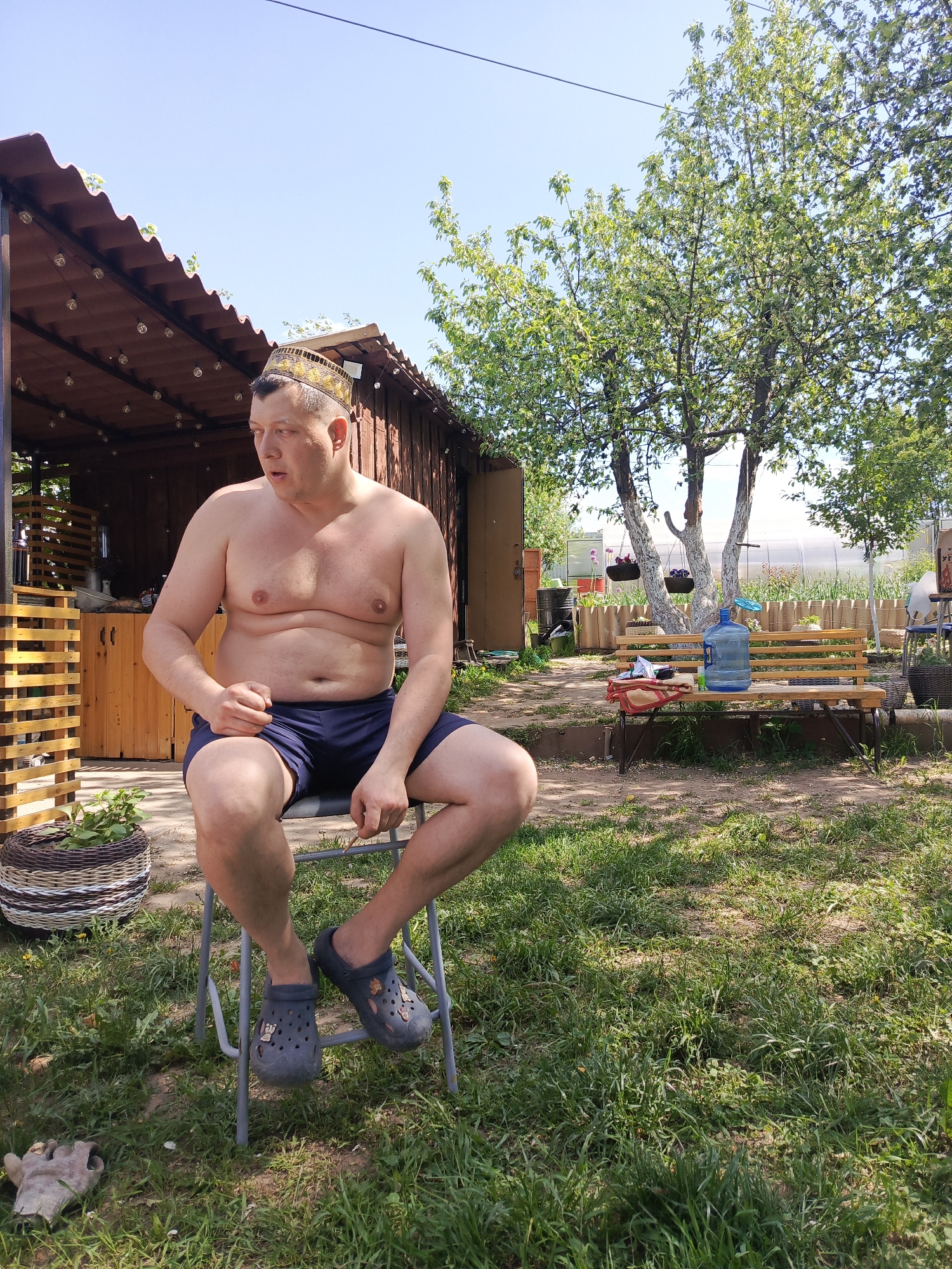 The image depicts a man seated on a gray metal folding chair in a yard in Chaikovsky, Russia. He is bare-chested, wearing navy blue athletic shorts, dark gray Crocs-style shoes, and a traditional Central Asian tubeteika cap. He is looking off to his left.
Behind him is a wooden structure with a corrugated metal roof and exposed light bulbs. Next to the chair is a woven basket containing foliage and a gardening glove on the ground.
In the background, there is a wooden bench with a large water jug on it. The bench is beneath a tree with whitewashed trunks and hanging planters. A greenhouse is visible further back, and the ground is covered with grass.