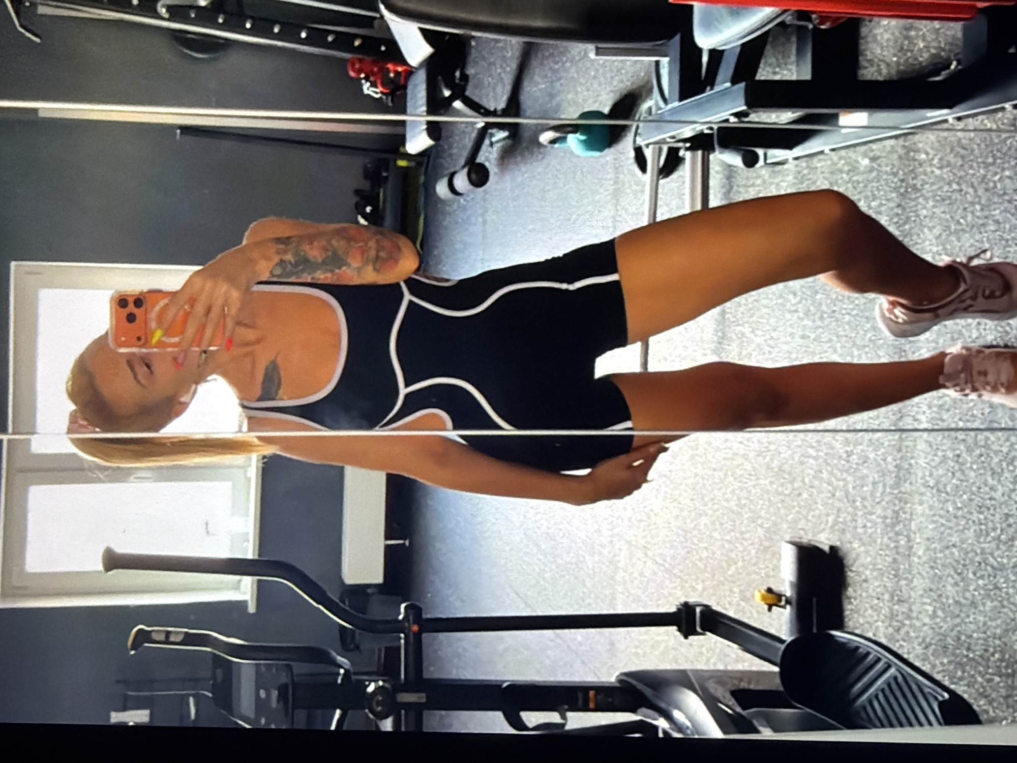 This image, rotated 90 degrees counter-clockwise, captures a woman taking a mirror selfie within what appears to be a gym or fitness studio.
The central subject is a woman, reflected from approximately the mid-thigh up, holding an orange-cased smartphone with her right hand to capture her reflection. Her face is partially obscured by the phone, which features a triple-lens camera system. She has long, light brown hair, possibly tied back, and a prominent, colorful tattoo sleeve covers her right arm. The woman is wearing a black one-piece workout suit with distinctive white wavy line trim across the bust and waist areas. Her athletic footwear, light pink and white sneakers, are visible on her feet.
The setting is an indoor environment with a speckled gray floor, commonly found in gyms. Various pieces of fitness equipment are reflected in the mirror. A sturdy black power rack or weight machine frame is visible in the upper background, along with a teal-colored kettlebell on the floor. In the foreground and lower section of the image, parts of an elliptical trainer or similar cardio machine, including its handlebars and pedals, are clearly visible. A window with a white frame is reflected in the background, suggesting external light, but the scene is predominantly lit by interior artificial lighting.
The activity taking place is the woman posing for and capturing a mirror selfie. Her posture and engagement with the phone indicate a self-portrait action, likely to showcase her workout attire or presence at the gym. There is no visible text in the image.