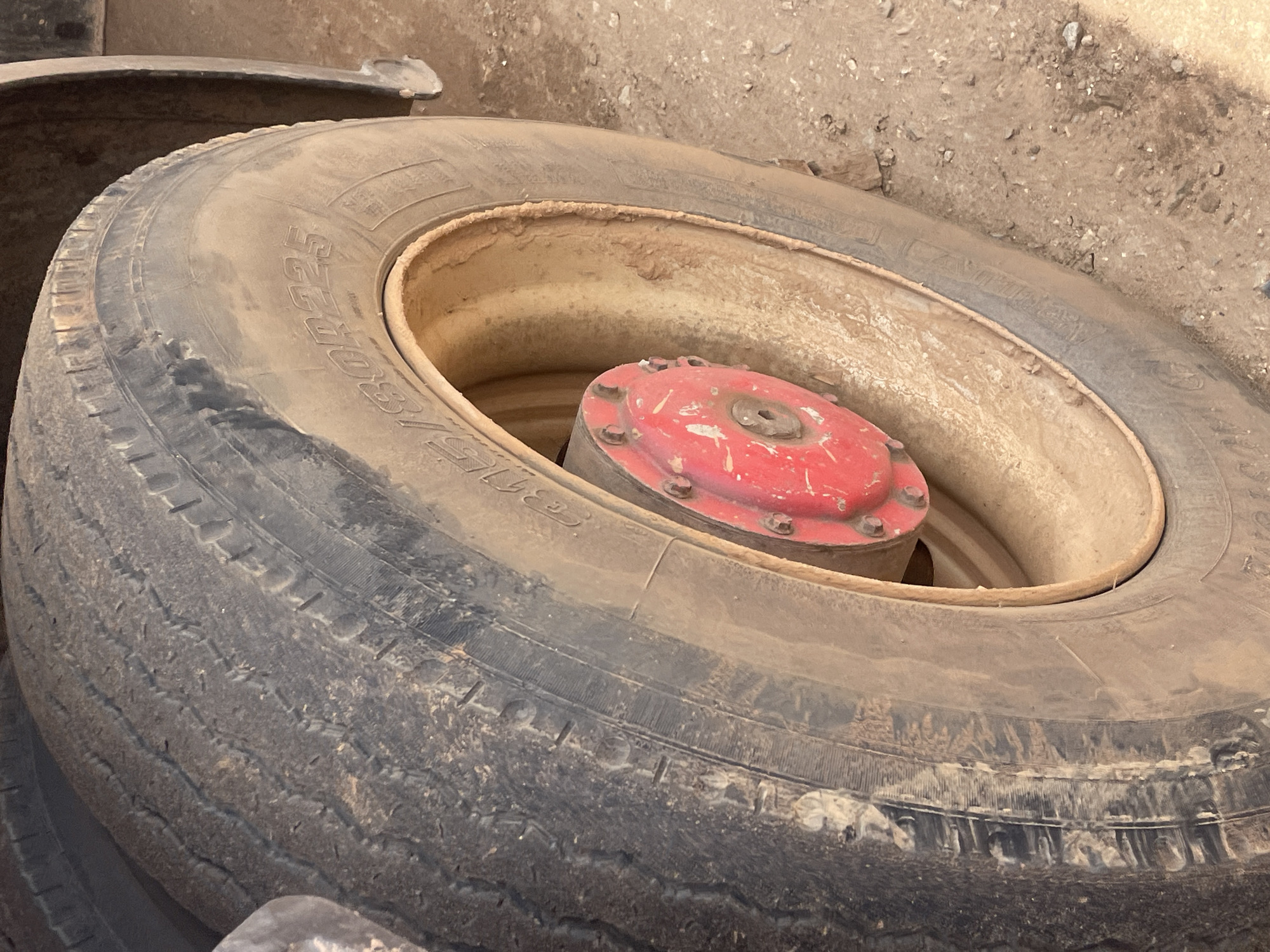 The image shows a close-up of a worn tire, likely from a vehicle such as a truck or tractor, resting on the ground. The tire is dirty and appears to have seen considerable use, with visible wear on the tread. Inside the rim of the tire, a red, circular hubcap with visible bolts is present. The background is a blurred expanse of dirt and small rocks, consistent with an outdoor, potentially rural or industrial setting. There are no people visible in the image. The lighting suggests it is daytime, but the weather is not discernible. Text is visible on the tire's sidewall, though it is partially obscured by dirt and the angle of the shot. The overall impression is one of a used, functional piece of equipment in a somewhat unkempt environment. The location context of Unguwar Saya, Nigeria, suggests this could be a scene related to transportation or agriculture in that region.