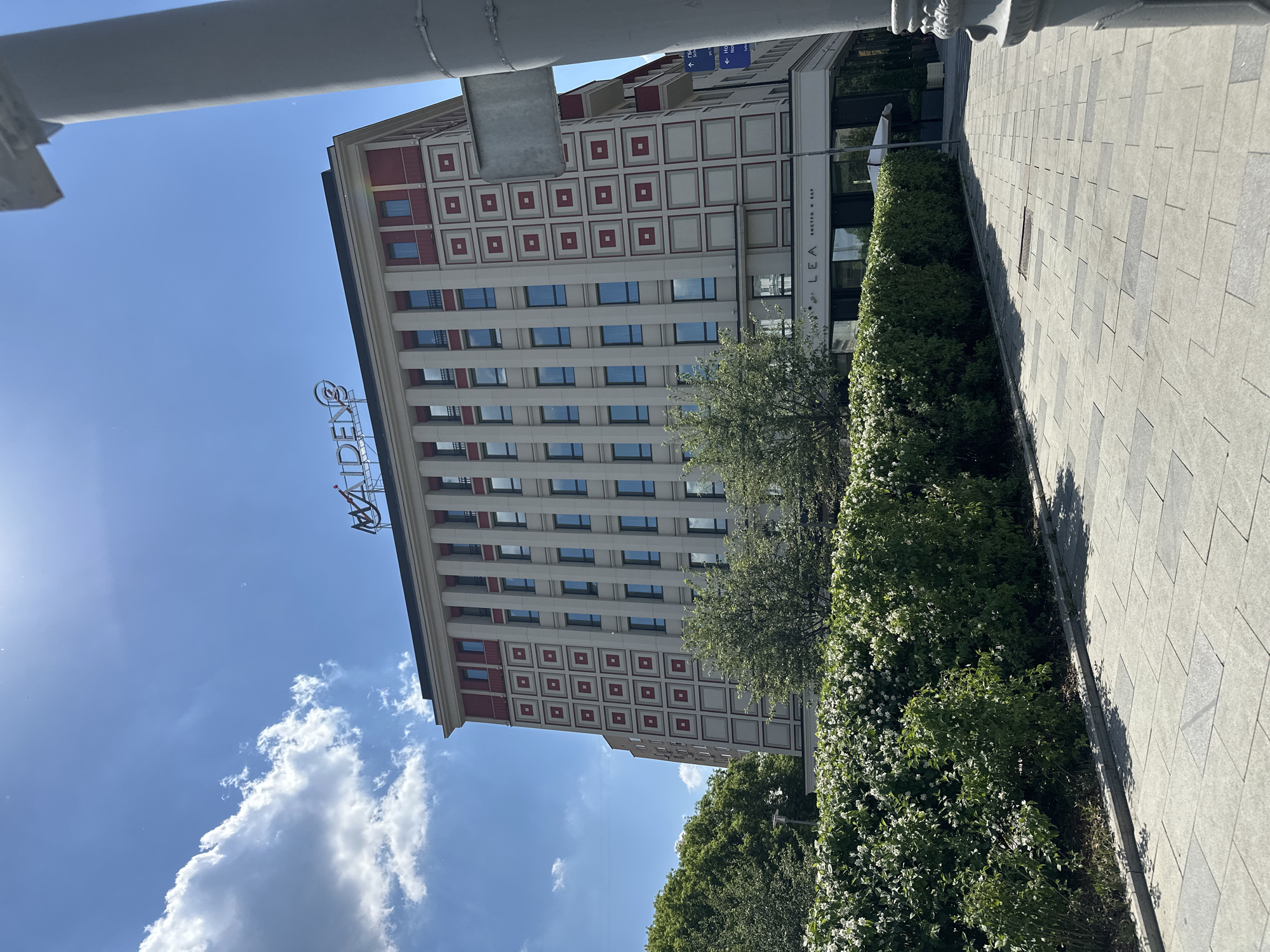 The image depicts the MAIDEN'S hotel building located in Moscow, Russia. The building has a distinct architectural style with a reddish-brown trim around the top and repeating square patterns on its facade. A neon sign reading "MAIDEN'S" is visible on the rooftop. In the foreground, a neatly trimmed hedge with white flowers runs along a paved sidewalk. The sky above is blue with scattered white clouds, and the sun is casting shadows. A streetlamp or column is partially visible on the right side of the frame. The ground is paved with large, light-colored rectangular tiles. The overall scene suggests a bright, sunny day in an urban setting.