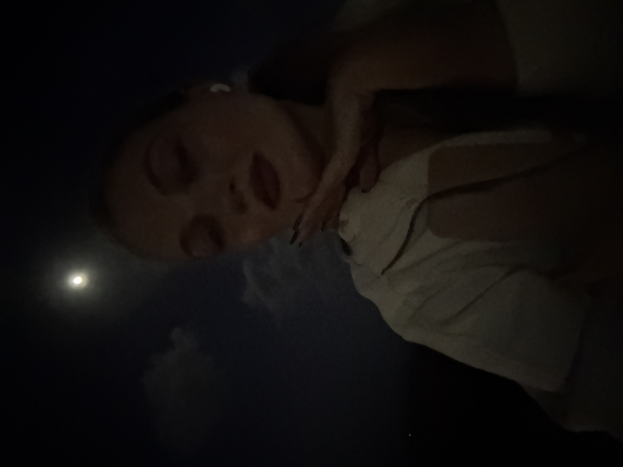 A young woman, with her face illuminated by the moon and some artificial light, is captured in a selfie at night. She is wearing a light-colored hooded jacket and has a hand resting on her chin. Her eyes are closed, and her lips are painted red. She is also wearing white earbuds. The background is a dark night sky with a bright moon and some clouds. The overall mood is serene and intimate. The image does not provide any specific details to identify its location in Denpasar, Indonesia.