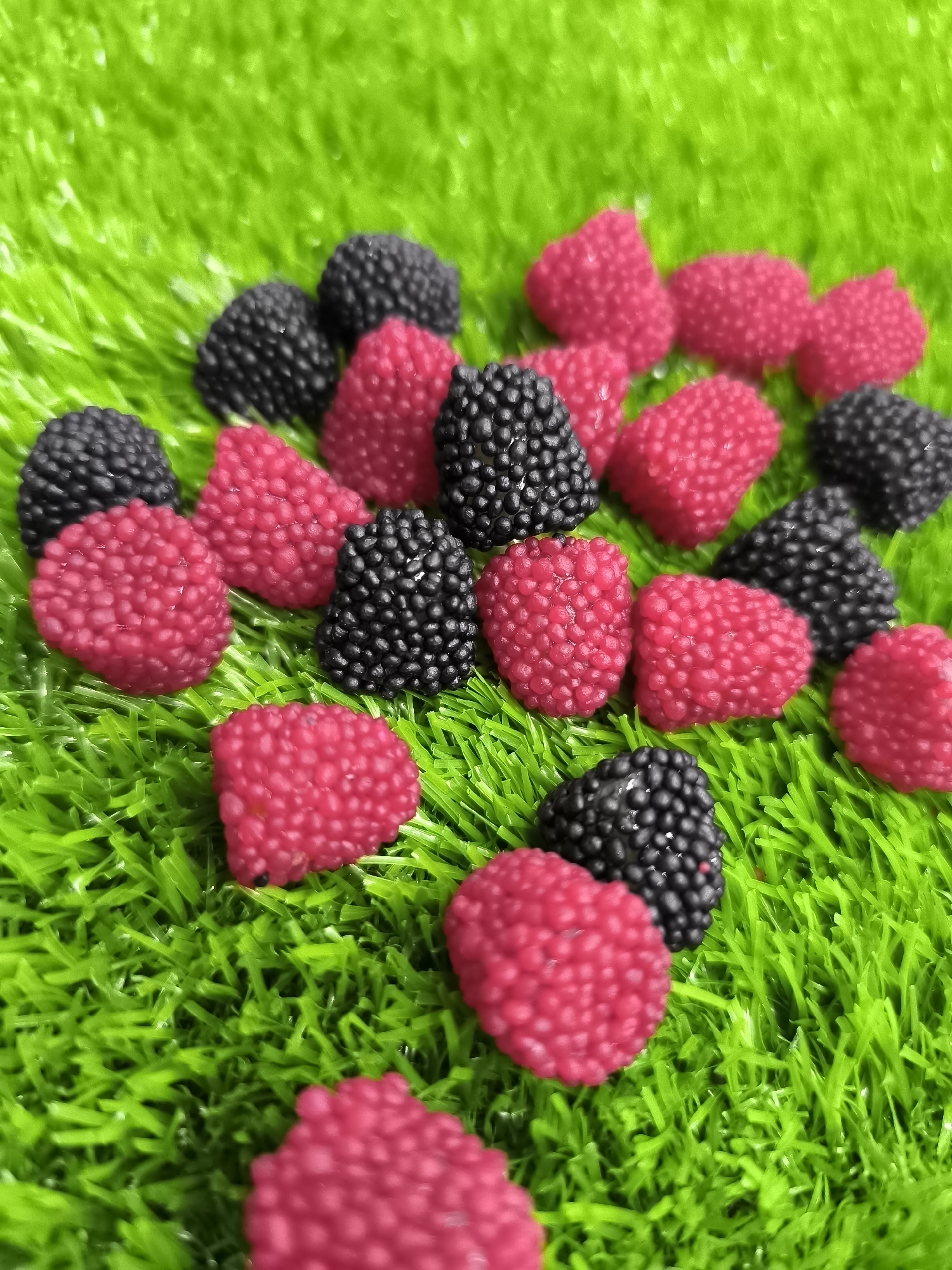 A top-down, medium shot captures a collection of artificial berries scattered across a bright green, artificial grassy surface. The berries are of two types: small, dark purple or black spheres clustered together, resembling blackberries, and larger, bright pink, bumpy spheres, resembling raspberries. These berries are arranged in a seemingly random pattern, with some overlapping and others scattered further apart. The artificial grass is densely packed and vibrantly green, creating a stark contrast with the berry colors. The lighting appears to be natural, casting subtle shadows that highlight the textured surfaces of the berries and the blades of grass. The scene is static, with no movement or human presence depicted. The image does not provide any information about the location of Kokand City, Uzbekistan.